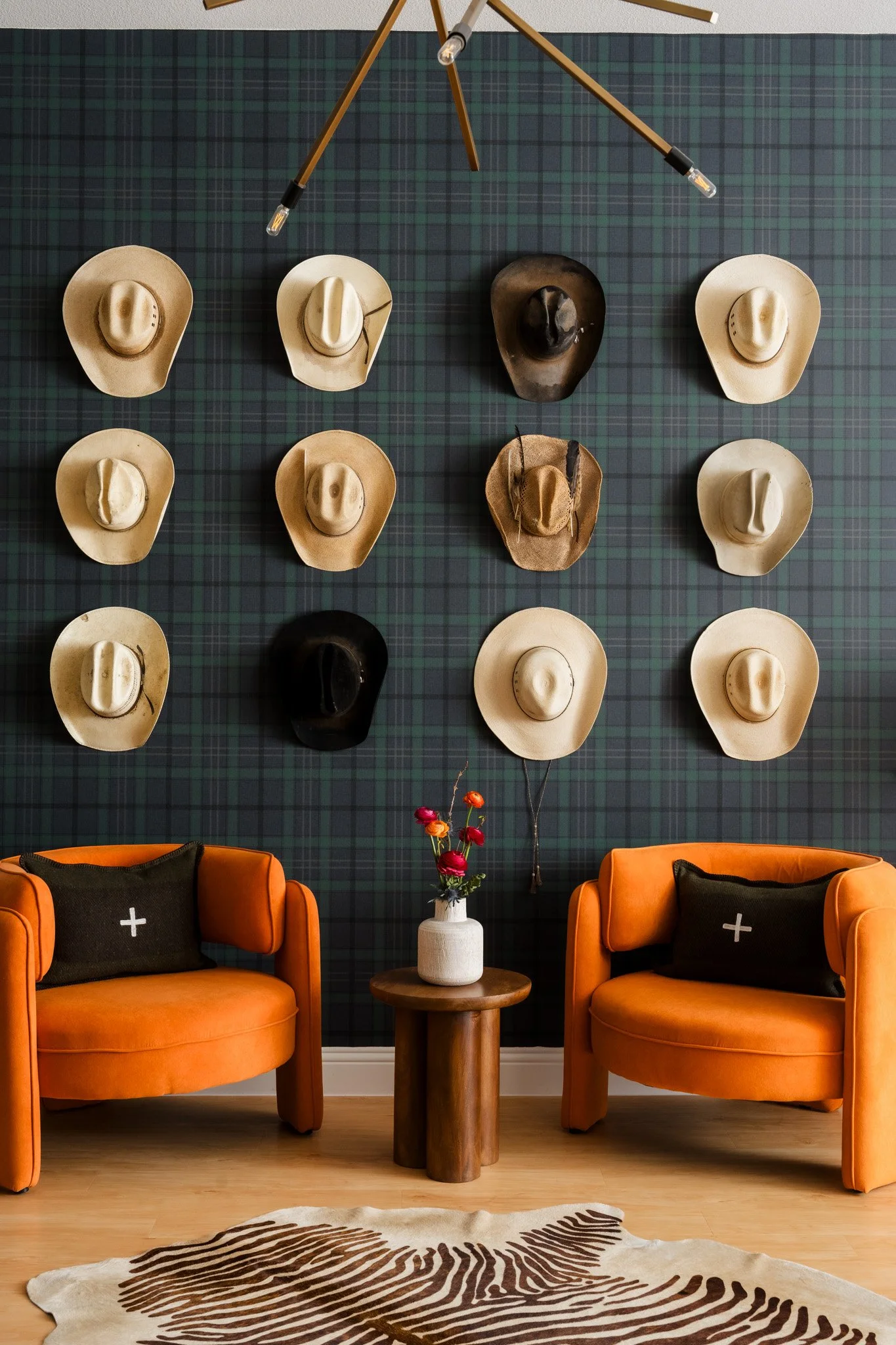 Texas commercial office Living room with two orange chairs, a small round wooden table with a white vase of flowers, a black and white animal-print rug, dark plaid wallpaper, and a wall display of cowboy hats in beige, black, and dark brown.