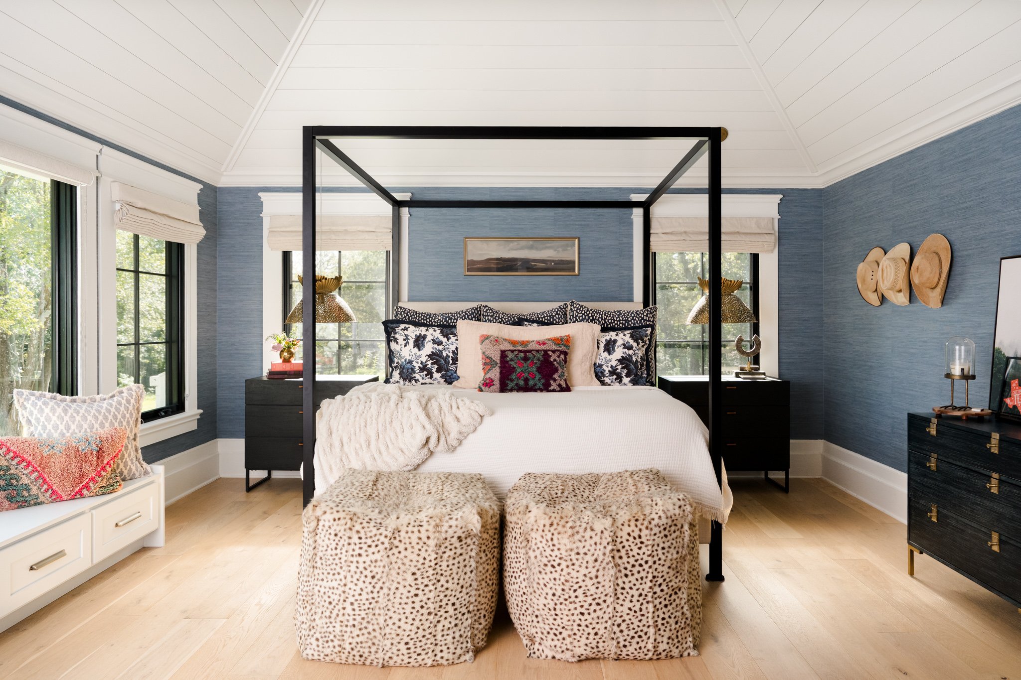 A bedroom with a four-poster bed, a blue textured wall, two windows with white shades, and various decorative pillows. Two leopard print ottomans are at the foot of the bed, and there are nightstands with lamps and decor on either side.