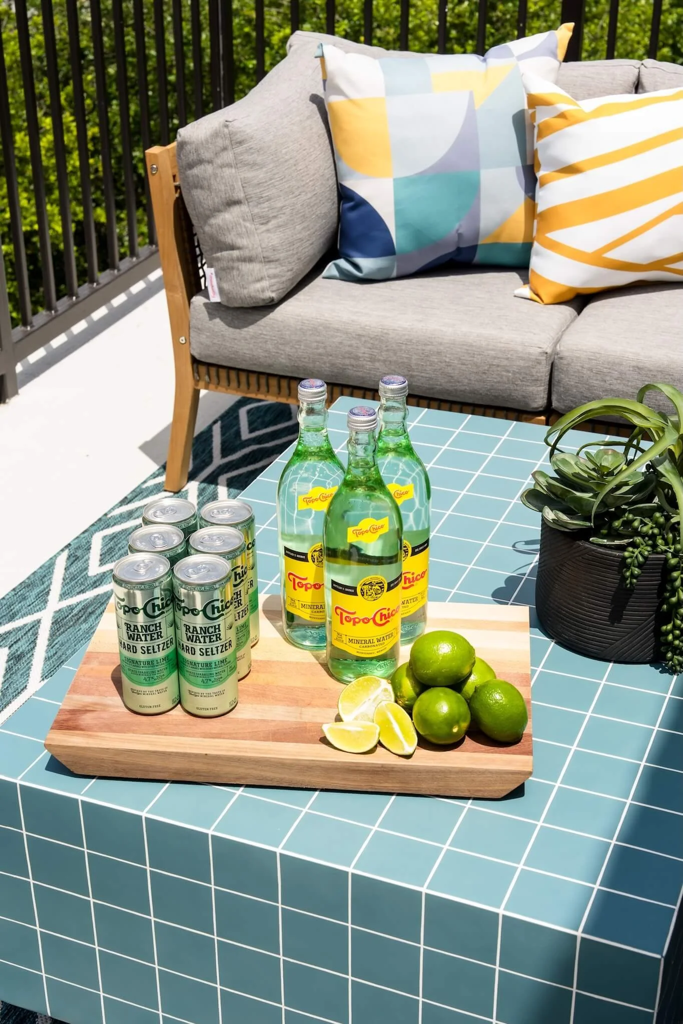 Outdoor patio table with four bottles of Topo Chico mineral water, six cans of Ranch Water hard seltzer, a wooden cutting board with fresh limes and lemon wedges, and a potted succulent plant on a blue and white checkered tablecloth. A gray sofa with
