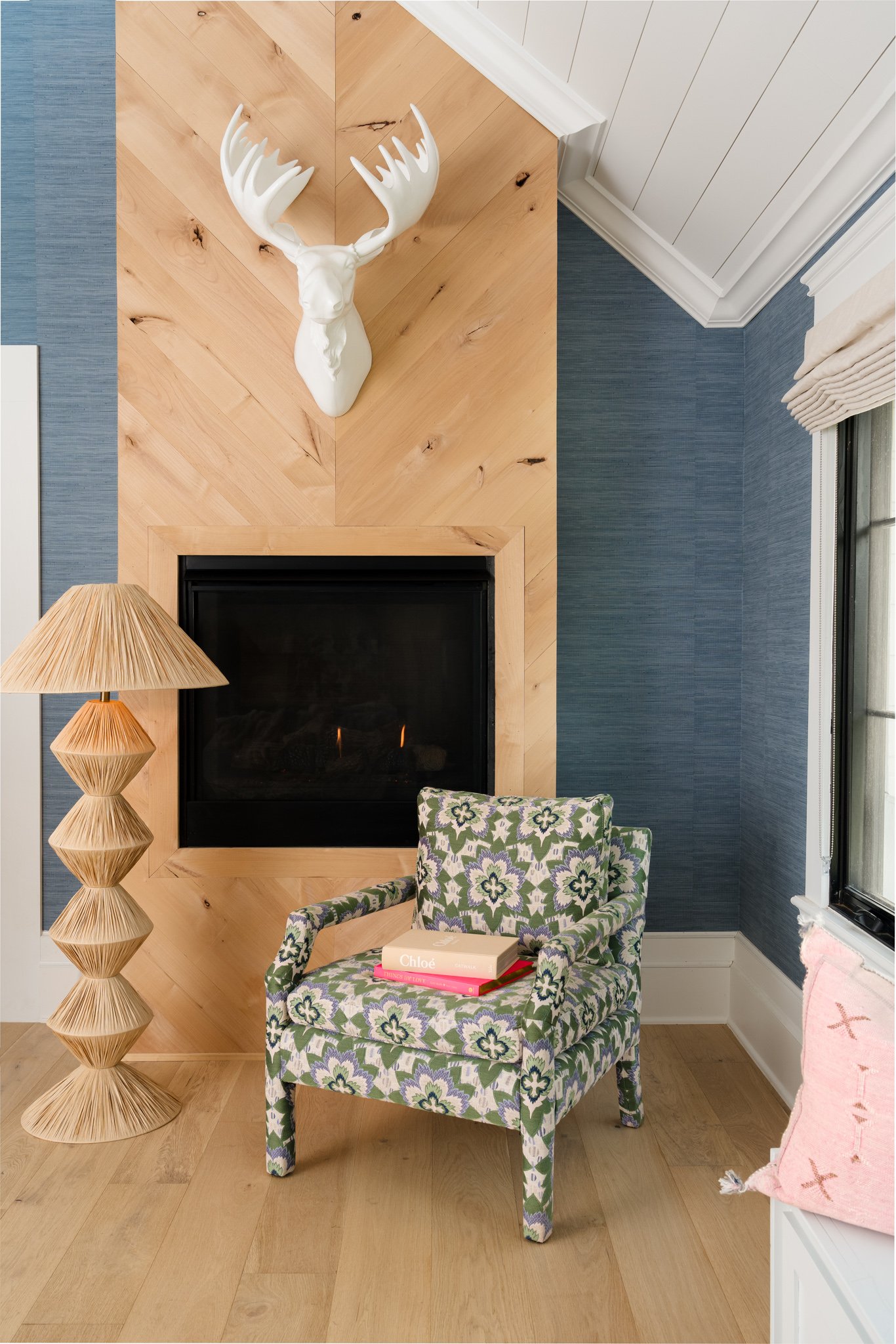 A living room corner with a patterned armchair, a tall wooden floor lamp, a white deer head wall decor, a fireplace, a blue-textured wall, a window with curtains, and a pink throw pillow.