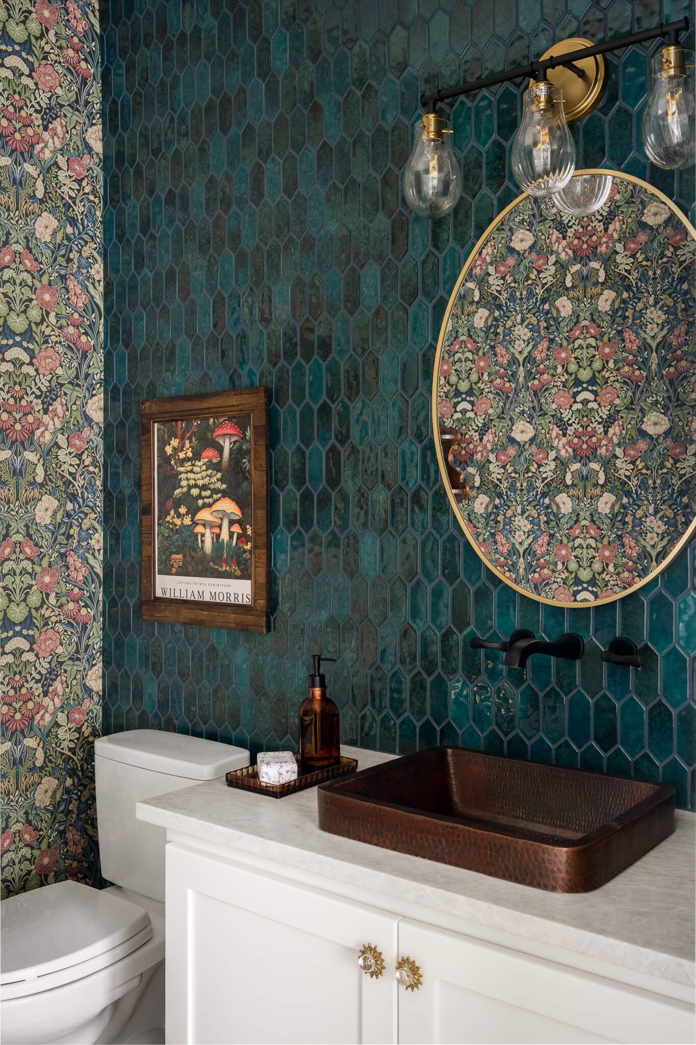 Decorative bathroom with dark teal hexagonal tile wall, floral wallpaper, oval mirror, vintage light fixture, white vanity, copper sink, and framed William Morris poster.