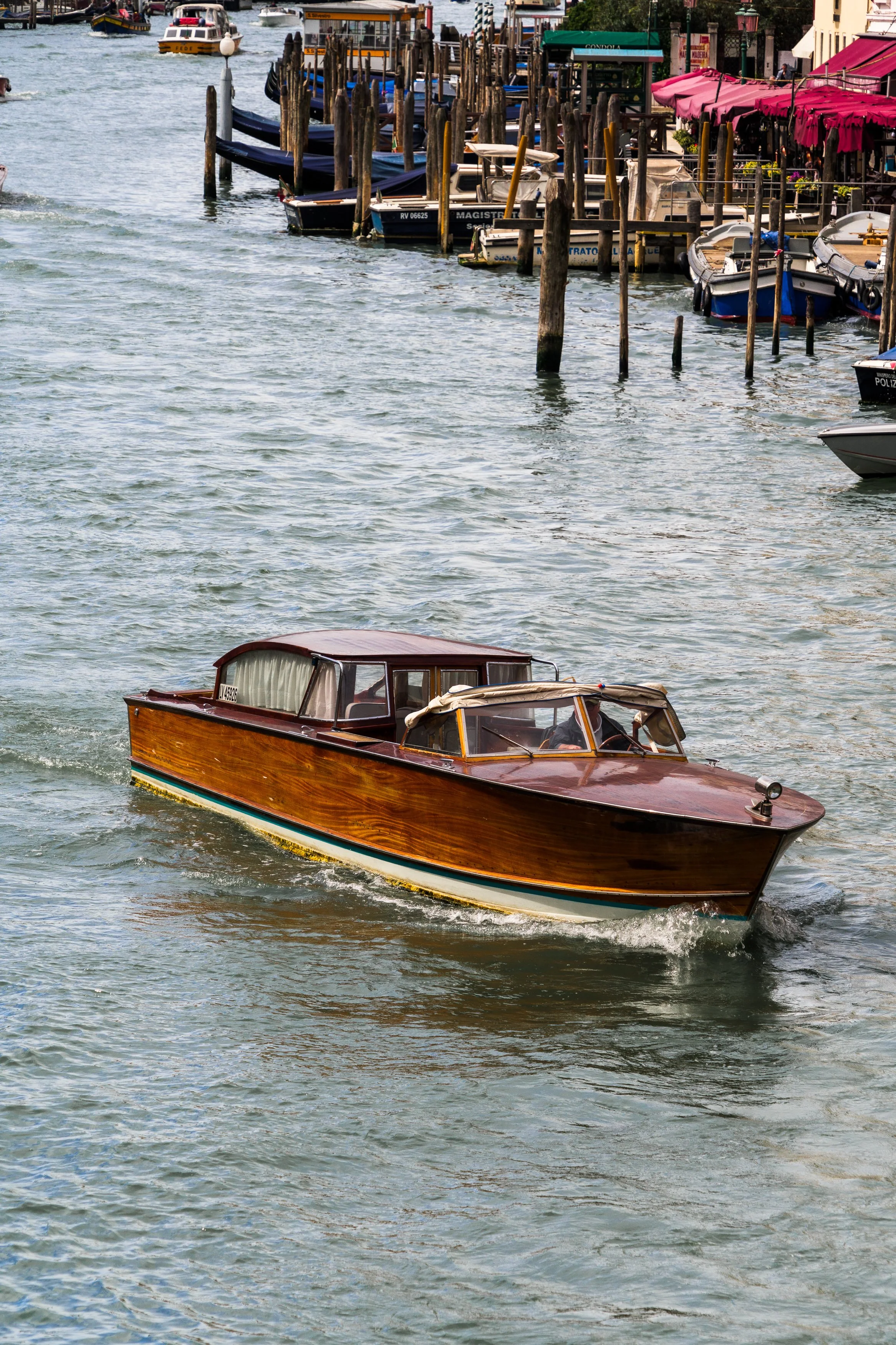Vintage wooden boat sailing on a canal with docks and boats on the side, colorful buildings in the background.