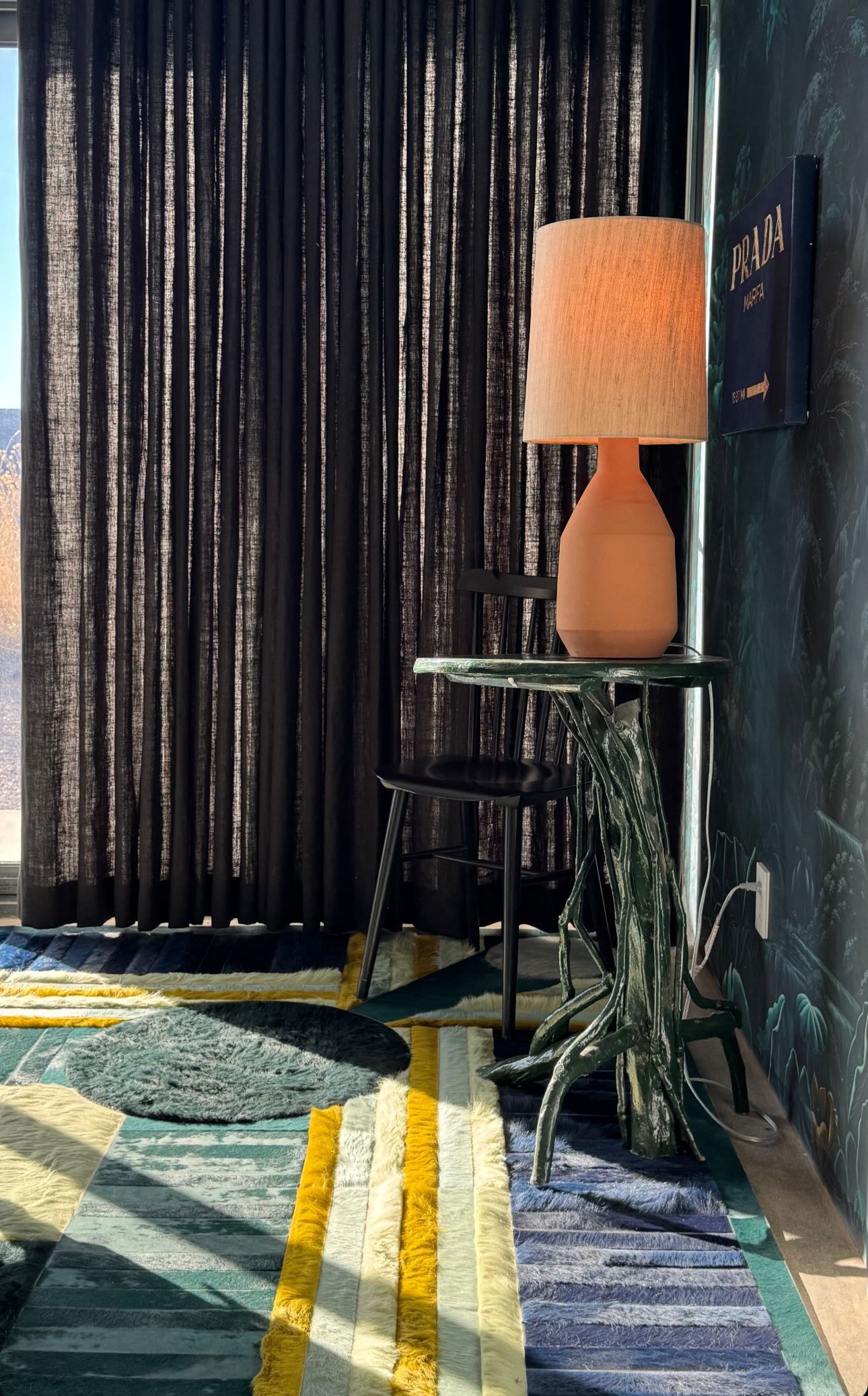 Interior of a room featuring black curtains, a green marble side table, a beige ceramic table lamp with a fabric lampshade, black chair, patterned walls, and multicolored geometric rugs on the floor.