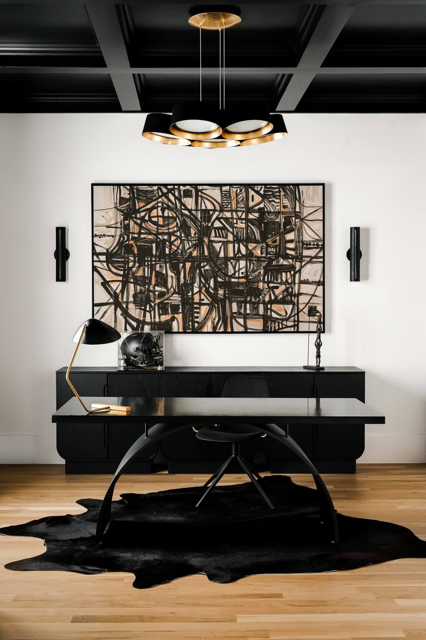 Modern black office desk with a black chair, decorative table lamp, and a cowhide rug in front. The background includes framed abstract art, a black cabinet, and black wall sconces.