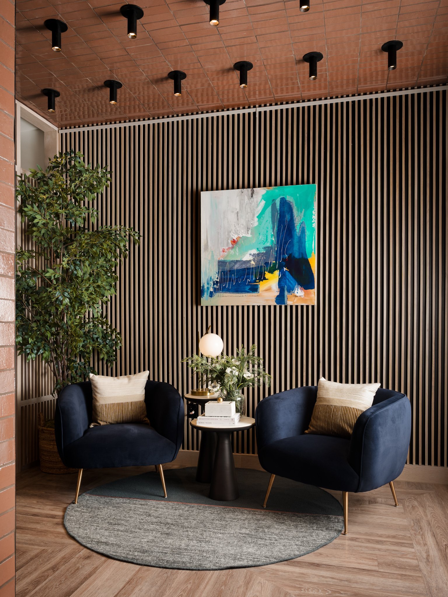 A cozy seating area with two dark blue armchairs, a round beige rug, a small black table with a lamp, books, and plants, set against a wooden slat wall with a colorful abstract painting. There are some pillows on the chairs and a large green leafy pl