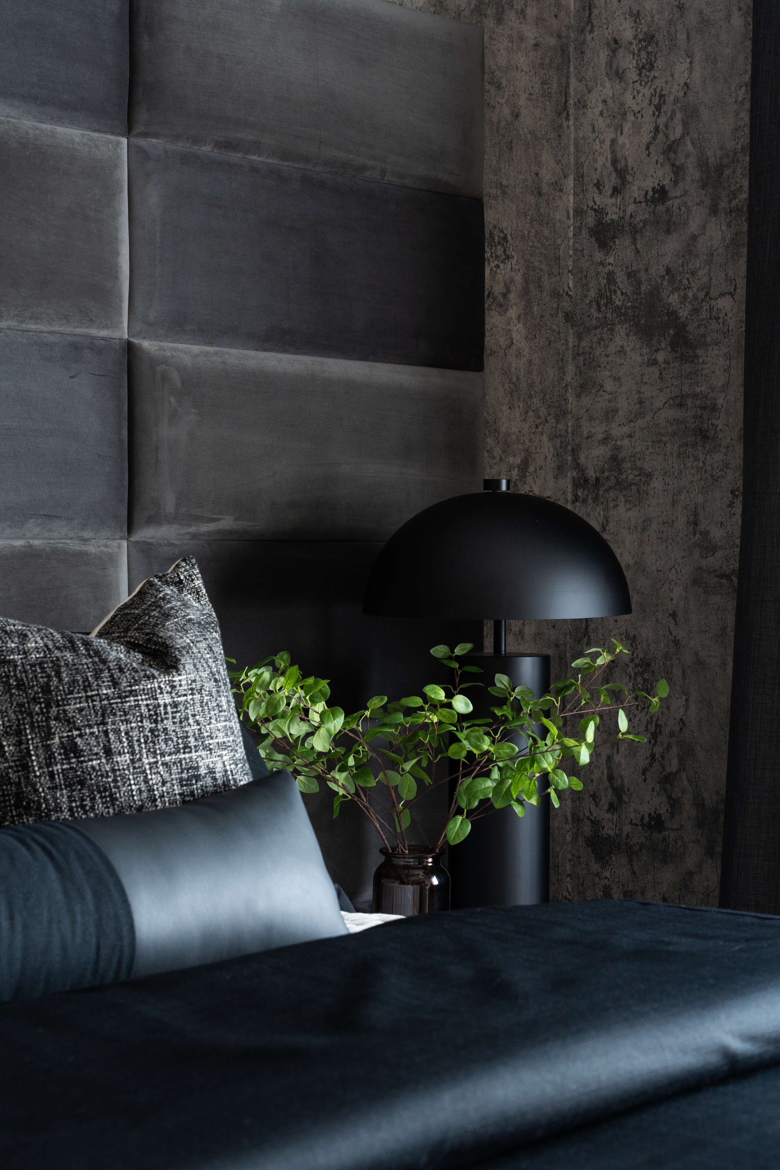 This dark and moody nightstand designed by Travis Pernell Interiors features a black metal and glass night stand, a black lamp, a custom velvet gray and black headboard. 