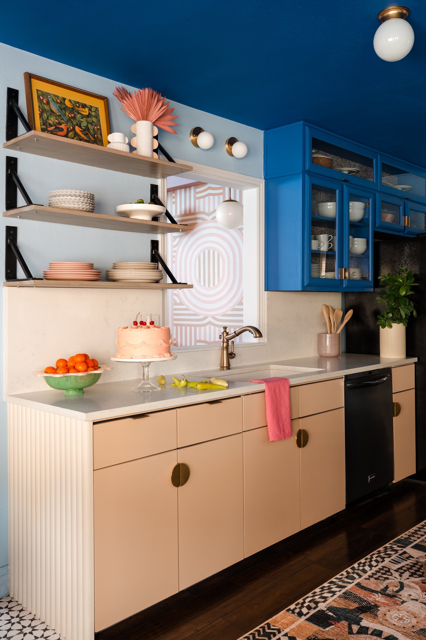 Modern kitchen with white cabinet, gold cabinet handles, black and white patterned floor, blue ceiling and upper cabinets, open shelving with dishes, fruit and cake on the countertop, pink napkin, and potted plant.