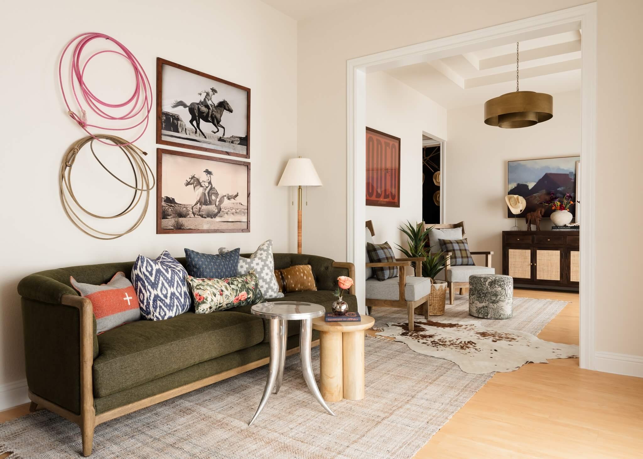 A cozy living room featuring a green sofa with colorful patterned pillows, a small silver side table, and wall art including black-and-white photographs of cowboys riding horses, along with woven basket and floral decor on a sideboard.