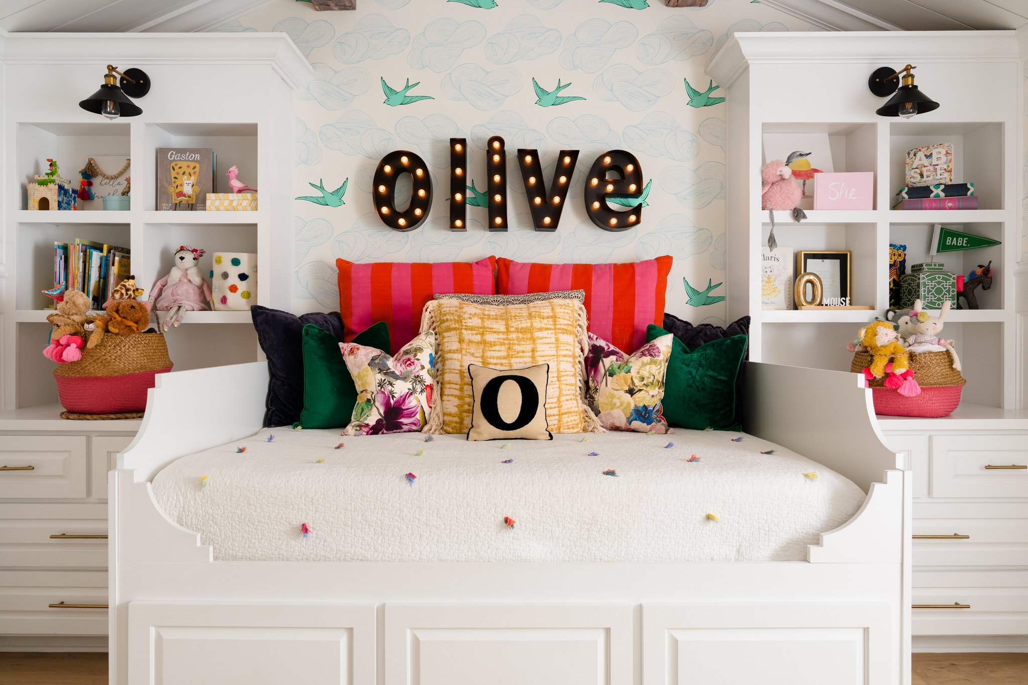 A cozy bedroom with two white bookshelves filled with toys, books, and decorations. A large white bed with colorful pillows and a string of small pom-poms on the blanket. Behind the bed is a wall with cloud and bird illustrations and a large marquee 
