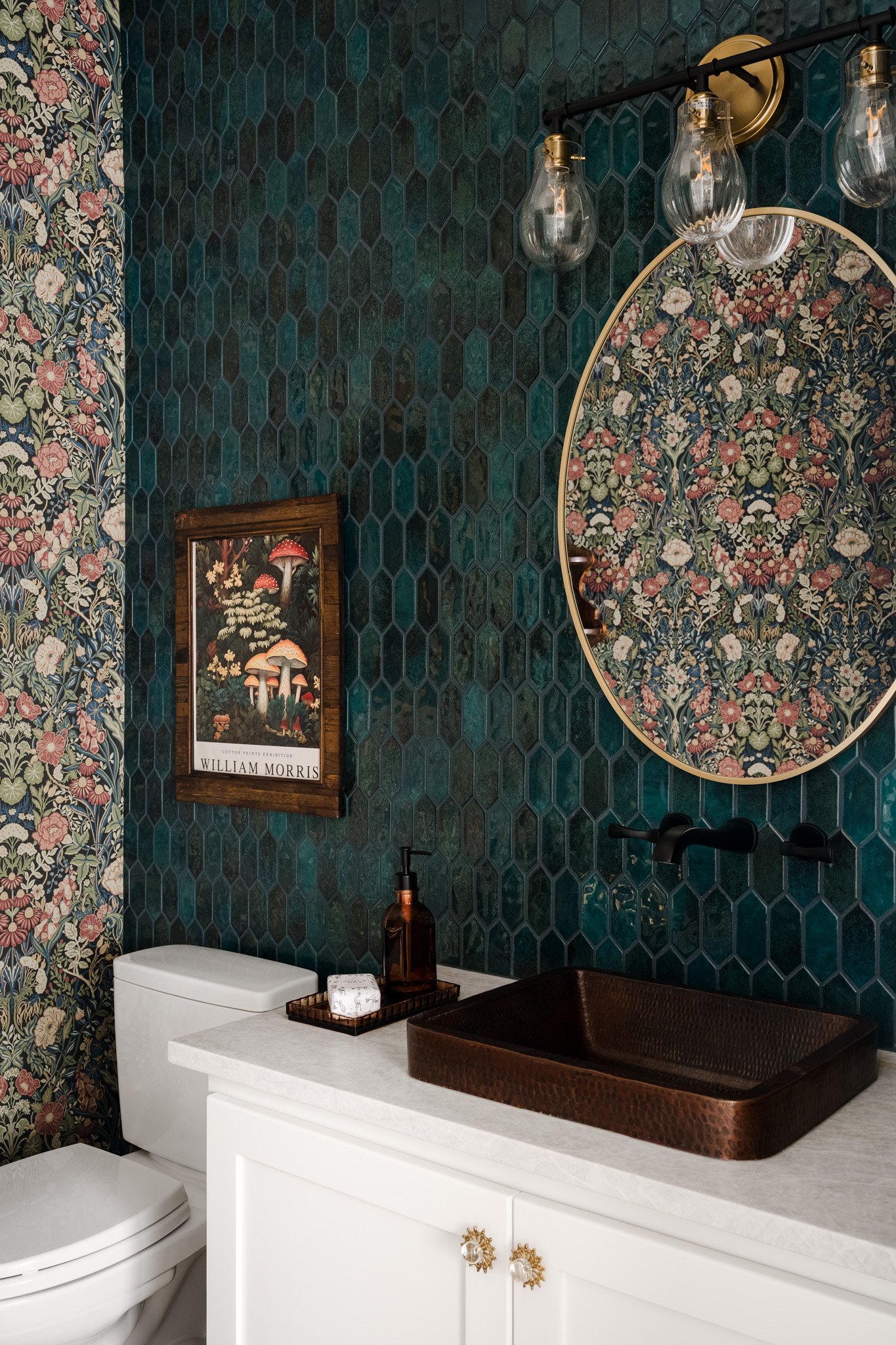 Bathroom with green hexagonal tile walls, floral-patterned mirror, framed mushroom print, toilet, and white vanity with decorative gold knobs. Woven brown tray on countertop holds soap dispenser and bar soap. Three glass bulb vanity lights above mirr