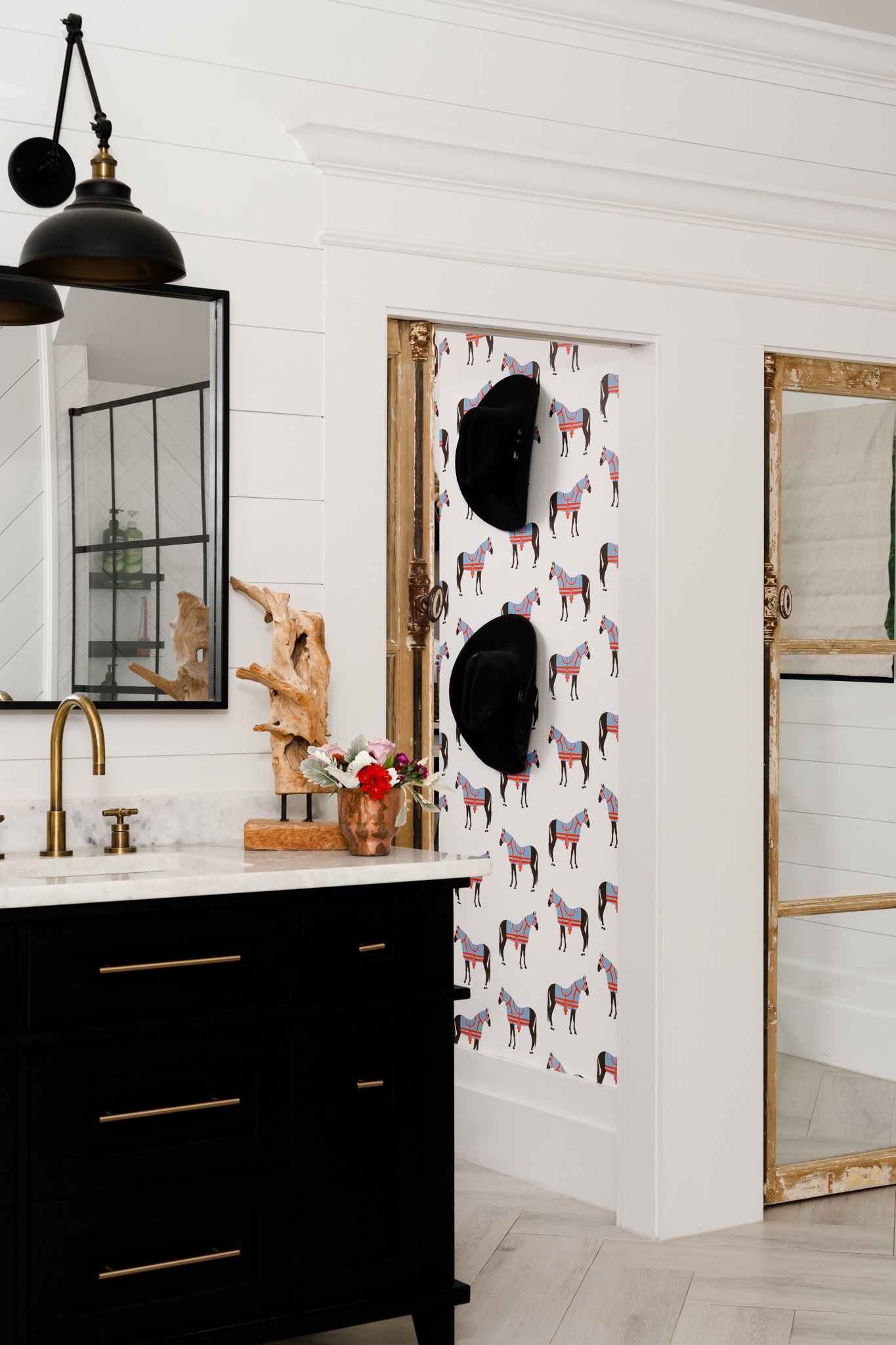 Indoor space with white shiplap walls, black pendant lights, a mirror, and a black cabinet with gold handles. Decorative elements include a wooden sculpture, a flower arrangement, and a wall with horse-patterned wallpaper. There are two doorframes, o
