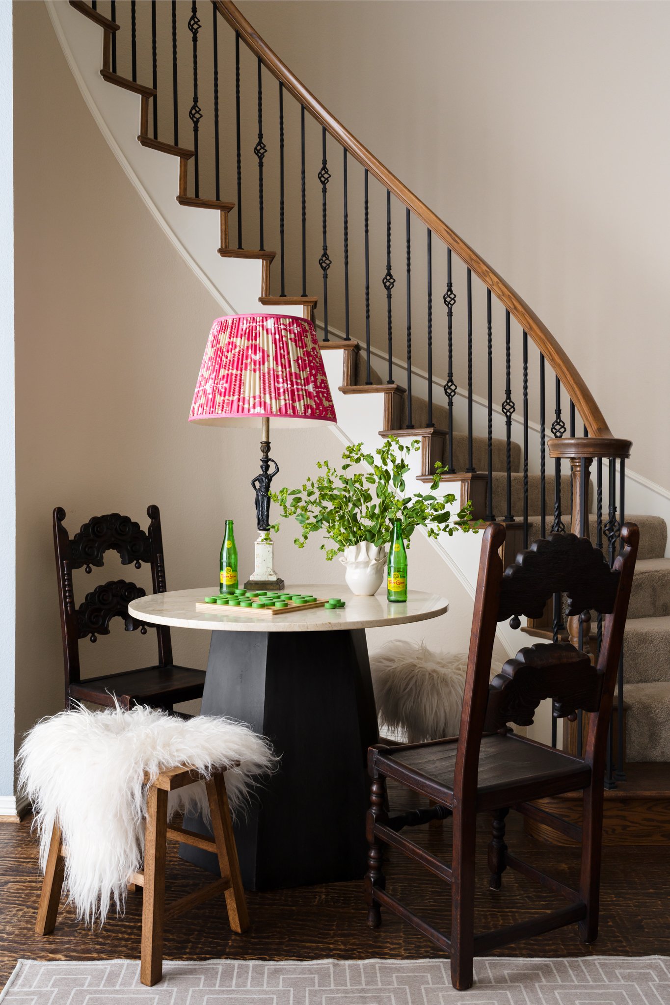 Interior scene of a home showing a staircase with a wooden handrail and black metal balusters, a dining table with dark wood chairs, a table lamp with a red patterned shade, a potted plant, two green glass bottles, white furry stools, and a beige wal