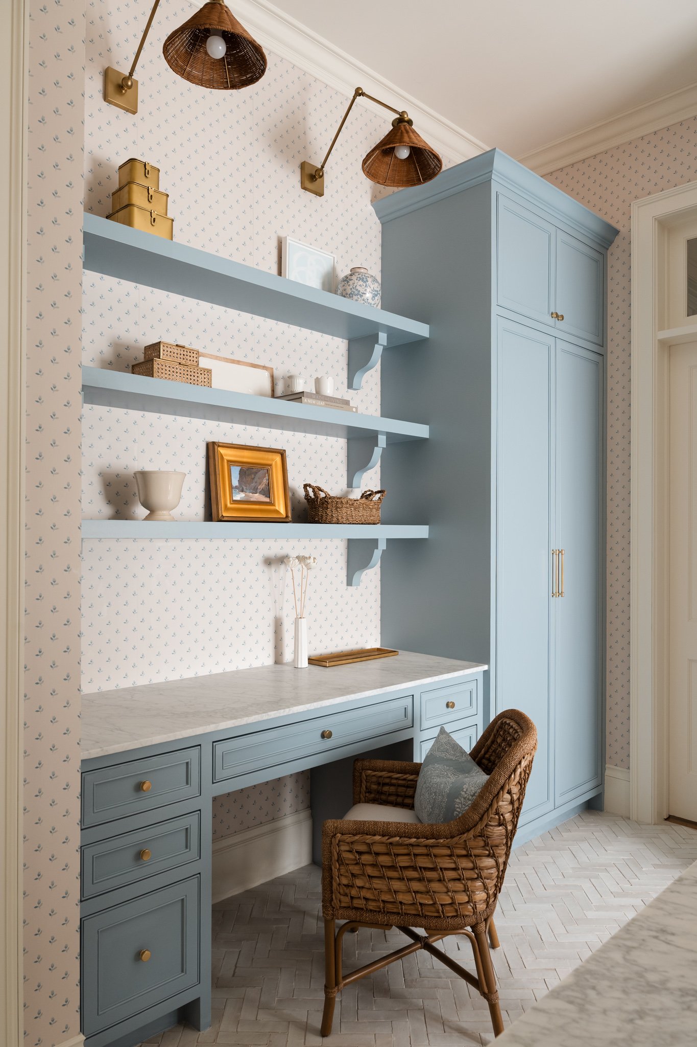 A cozy home workspace with a blue built-in desk and shelving unit, wicker chair with cushion, patterned wallpaper, and decorative items including a gold frame, small boxes, a bowl, and a lamp with two light fixtures.