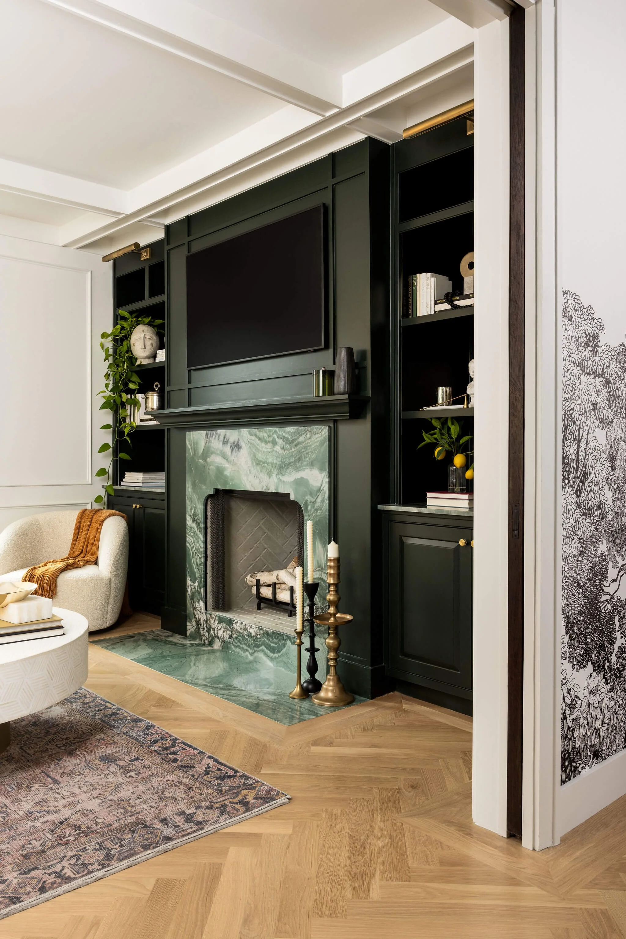 Living room corner featuring a dark green built-in bookshelf and fireplace with a green marble surround, a large flat-screen TV above, decorative candles on brass and black candlestick holders, a beige armchair with a rust-colored throw, a patterned 