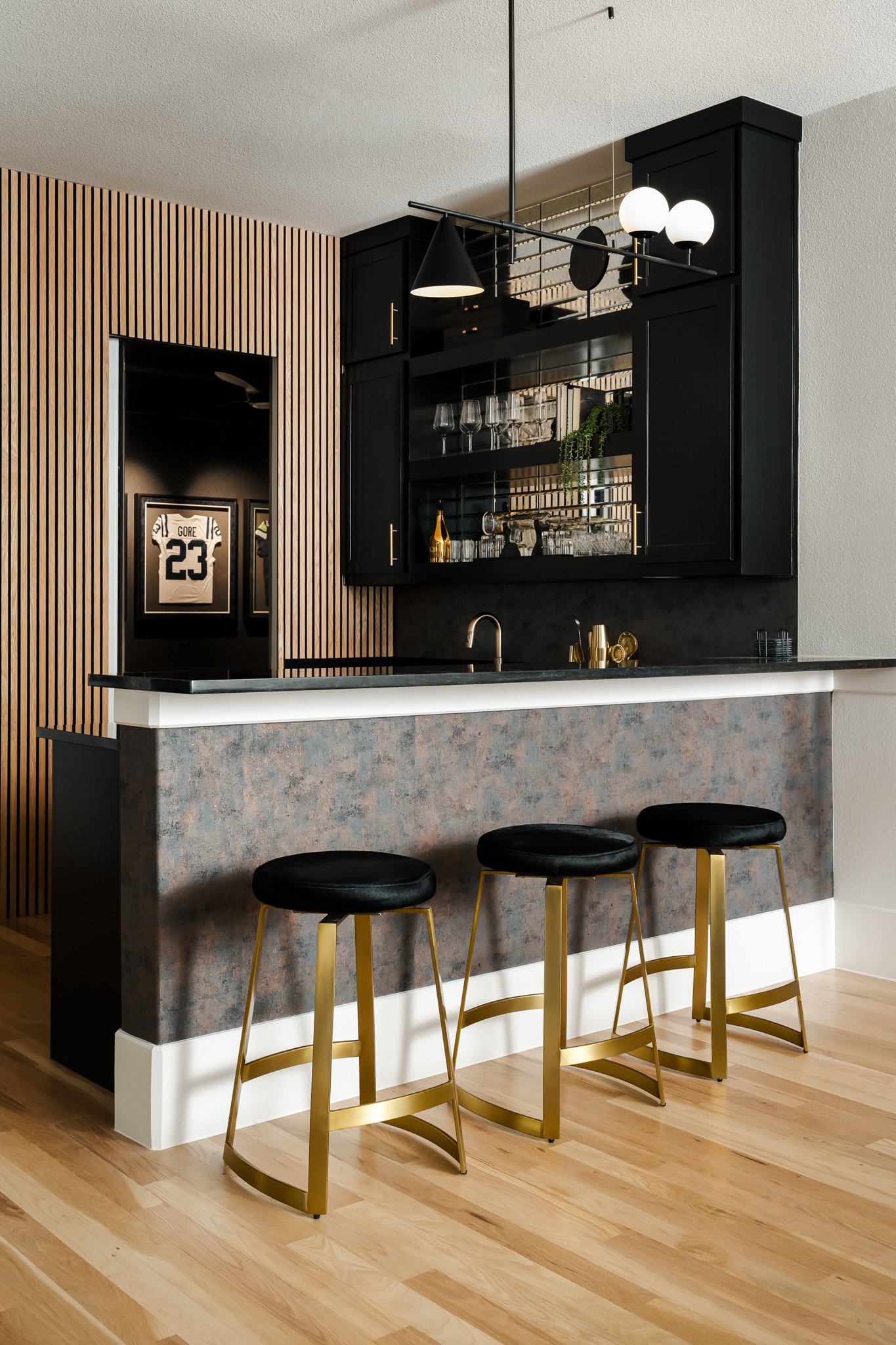 Modern kitchen bar area with four gold bar stools with black cushions, black cabinets with glassware, and a wood-paneled wall.