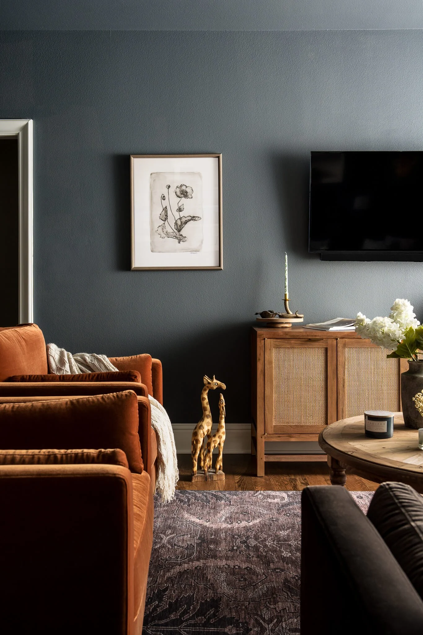 Living room with dark blue wall, framed black-and-white botanical art, flat-screen TV, wooden sideboard with candlestick, white flowers in a vase, two giraffe statues, orange velvet sofa, patterned area rug, and a round wooden coffee table.
