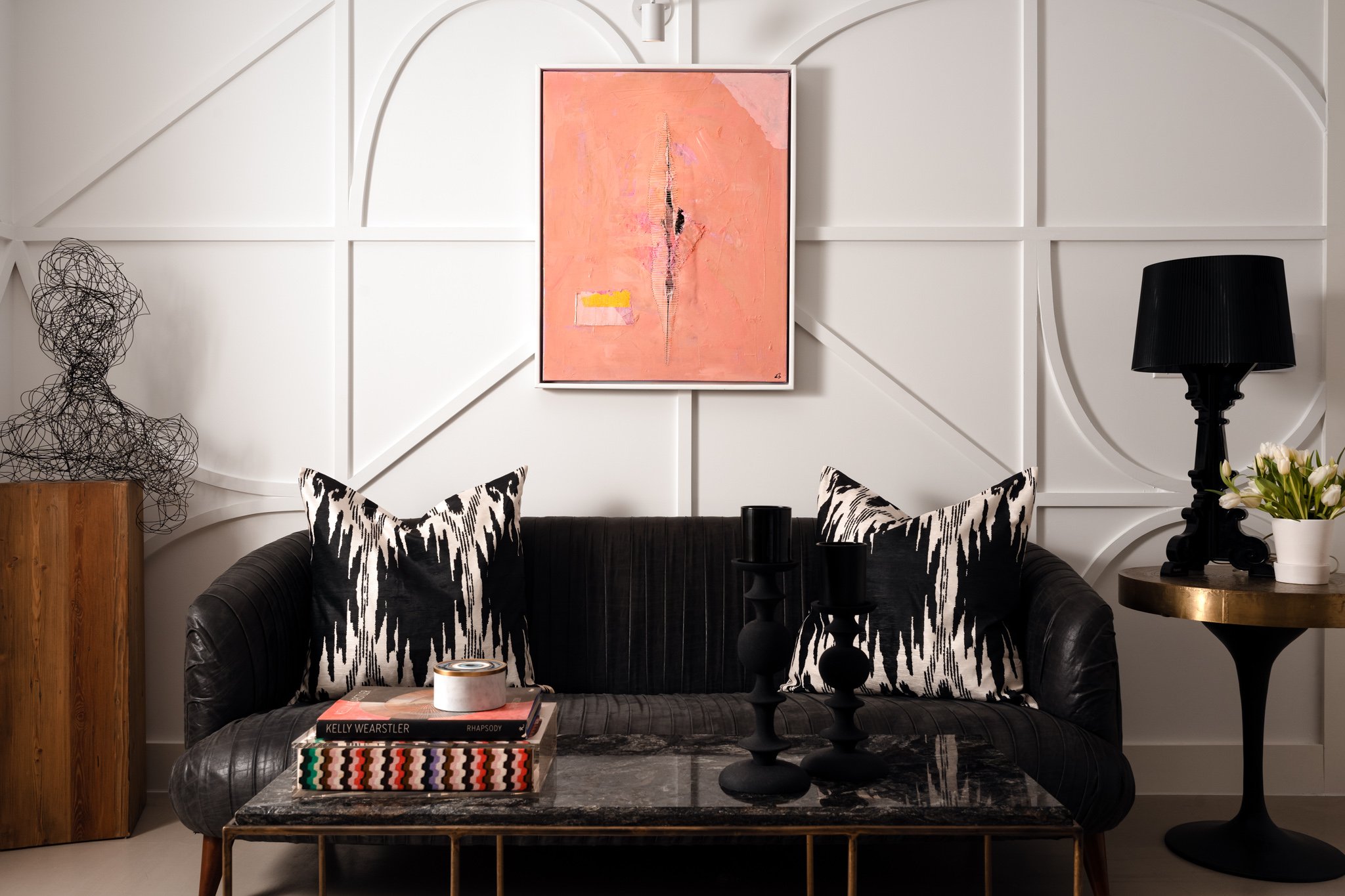 Living room with black leather sofa, zebra print pillows, abstract pink artwork on white paneled wall, black table with candles, marble coffee table with books and a colorful woven box, black floor lamp, flowers in white vase, wooden side table with 