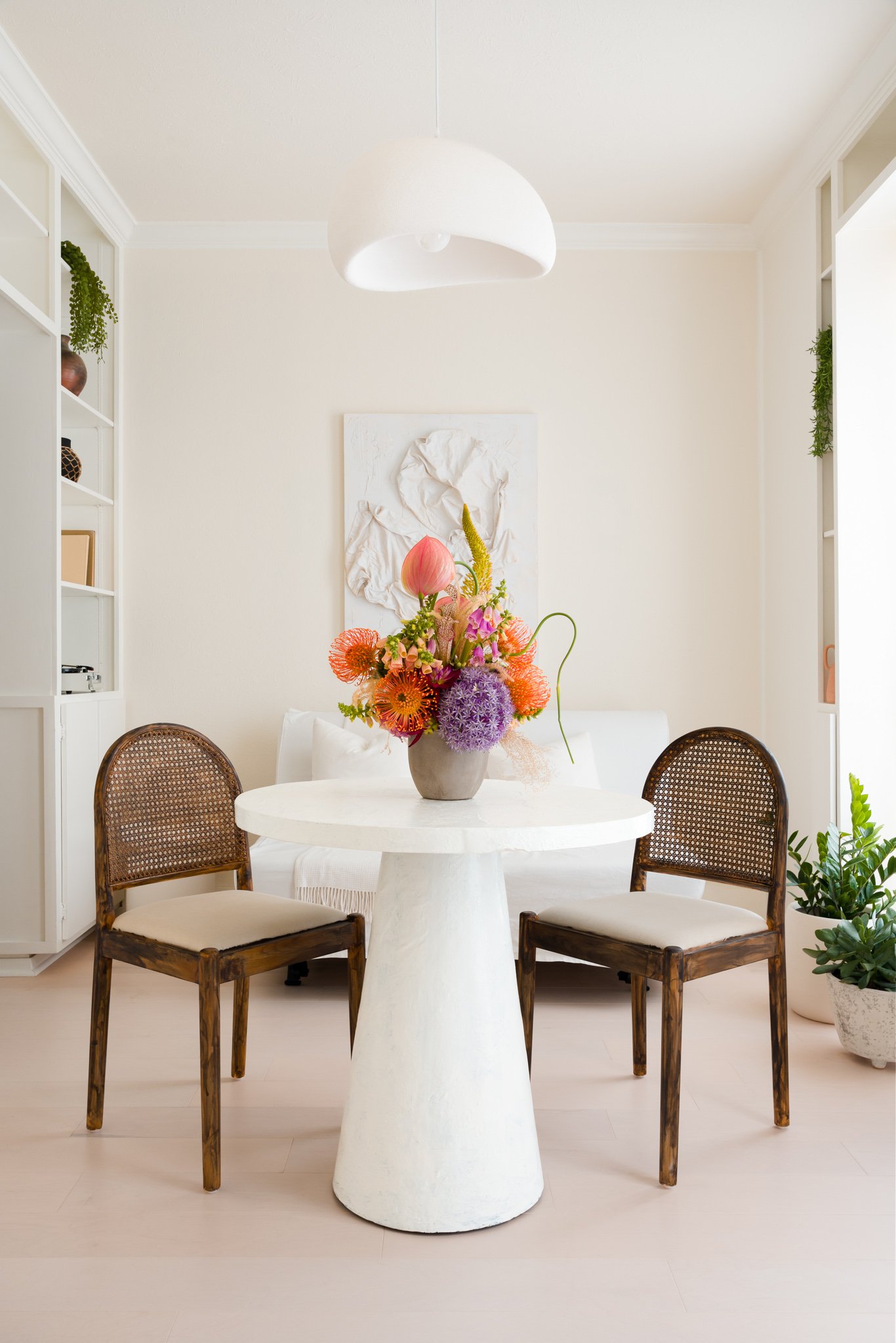 A round white dining table topped with a colorful flower arrangement in a gray vase, surrounded by four wooden chairs with woven cane backs and white cushions, in a bright, modern dining area with built-in white shelves and green plants.