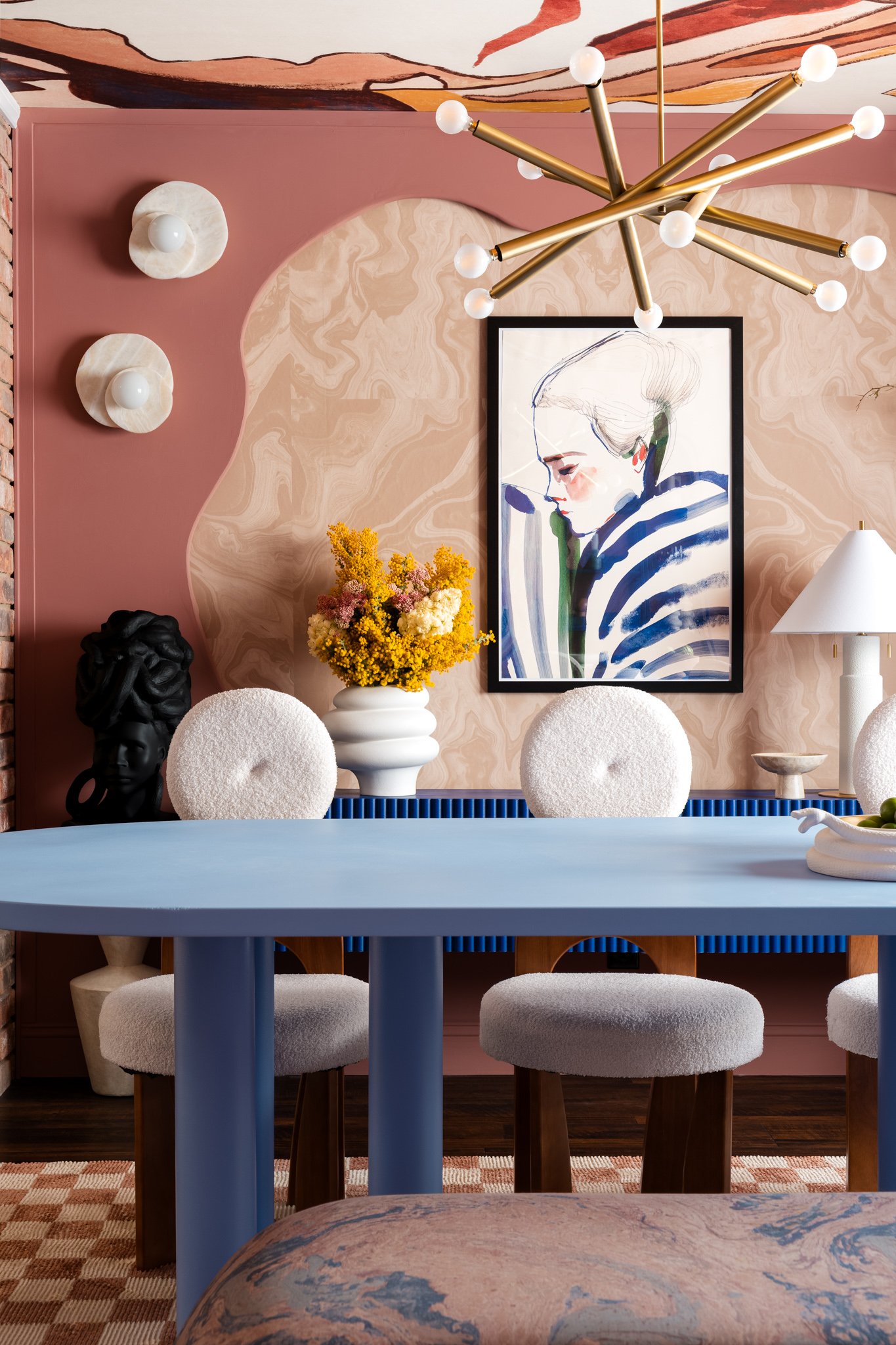 A dining room with a pink and marble textured wall, a modern gold chandelier, a framed line art portrait of a woman, a vase of yellow and purple flowers, white upholstered chairs around a blue table, and decorative wall sconces.