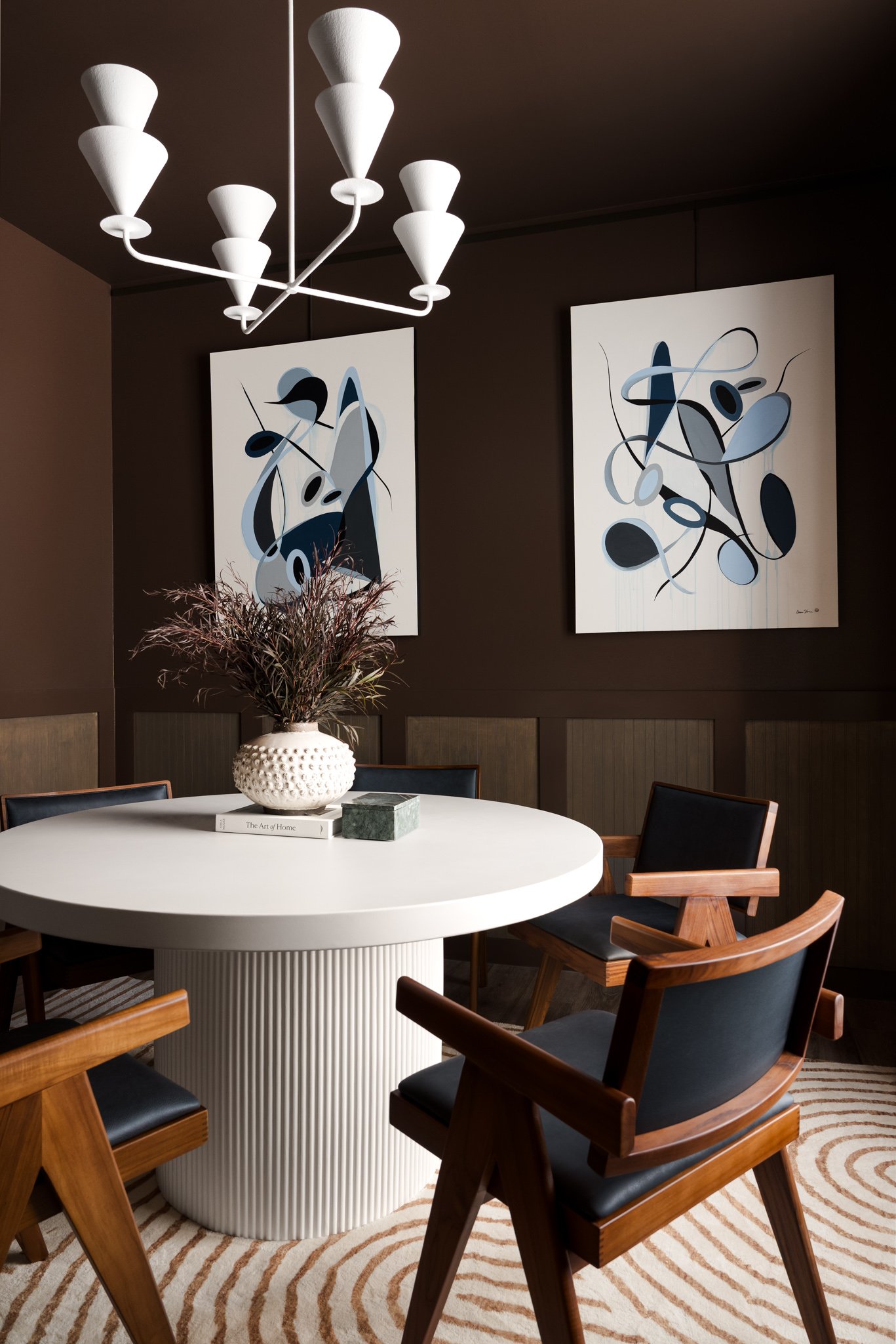 A dining room with a white round table, wooden chairs with black cushions, a white textured vase with dried plants, abstract artwork on the walls, a white chandelier, and a patterned rug.