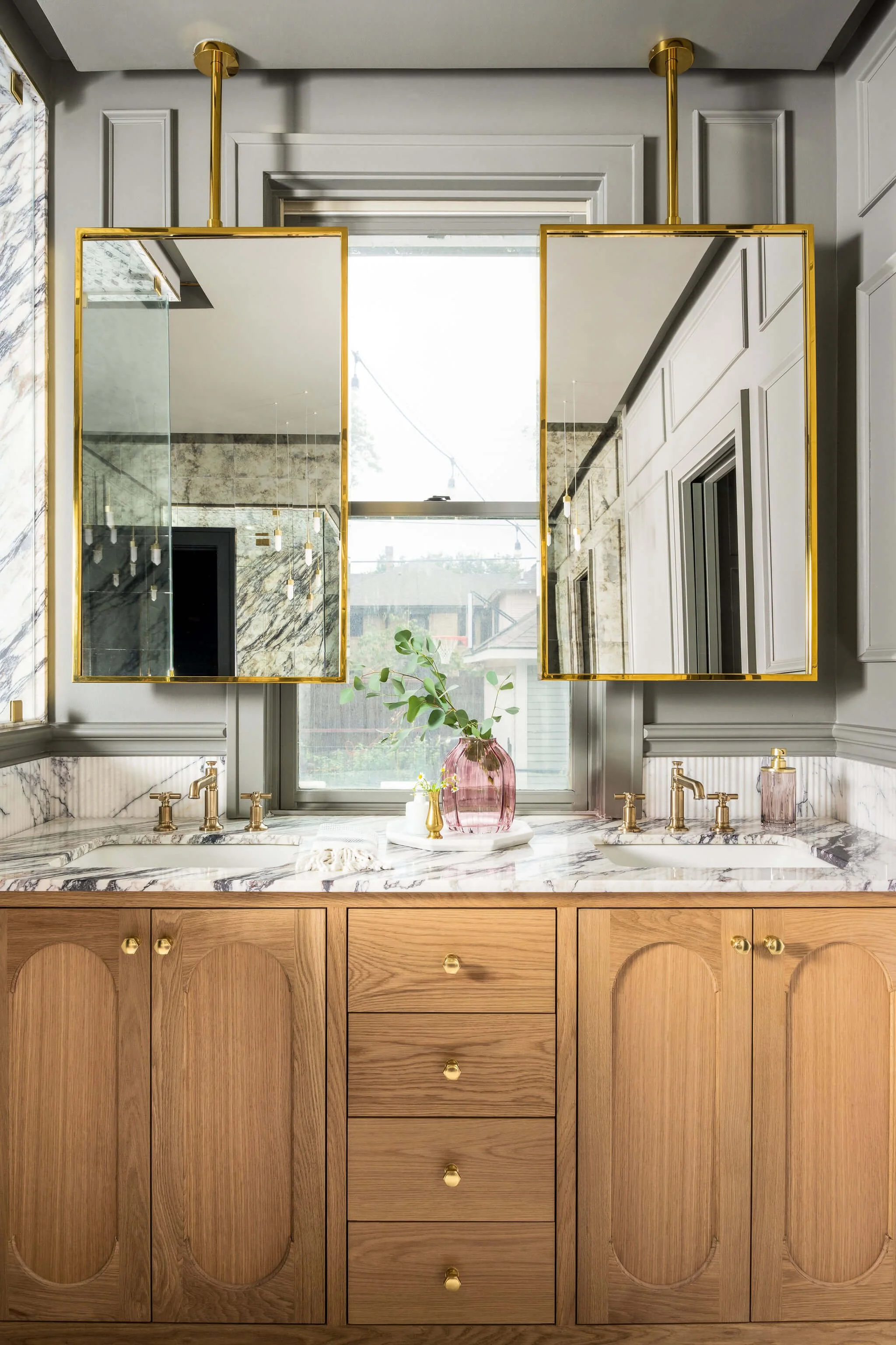 Bathroom vanity with marble countertop, gold fixtures, a pink vase with green foliage, and two mirrored medicine cabinets above a window