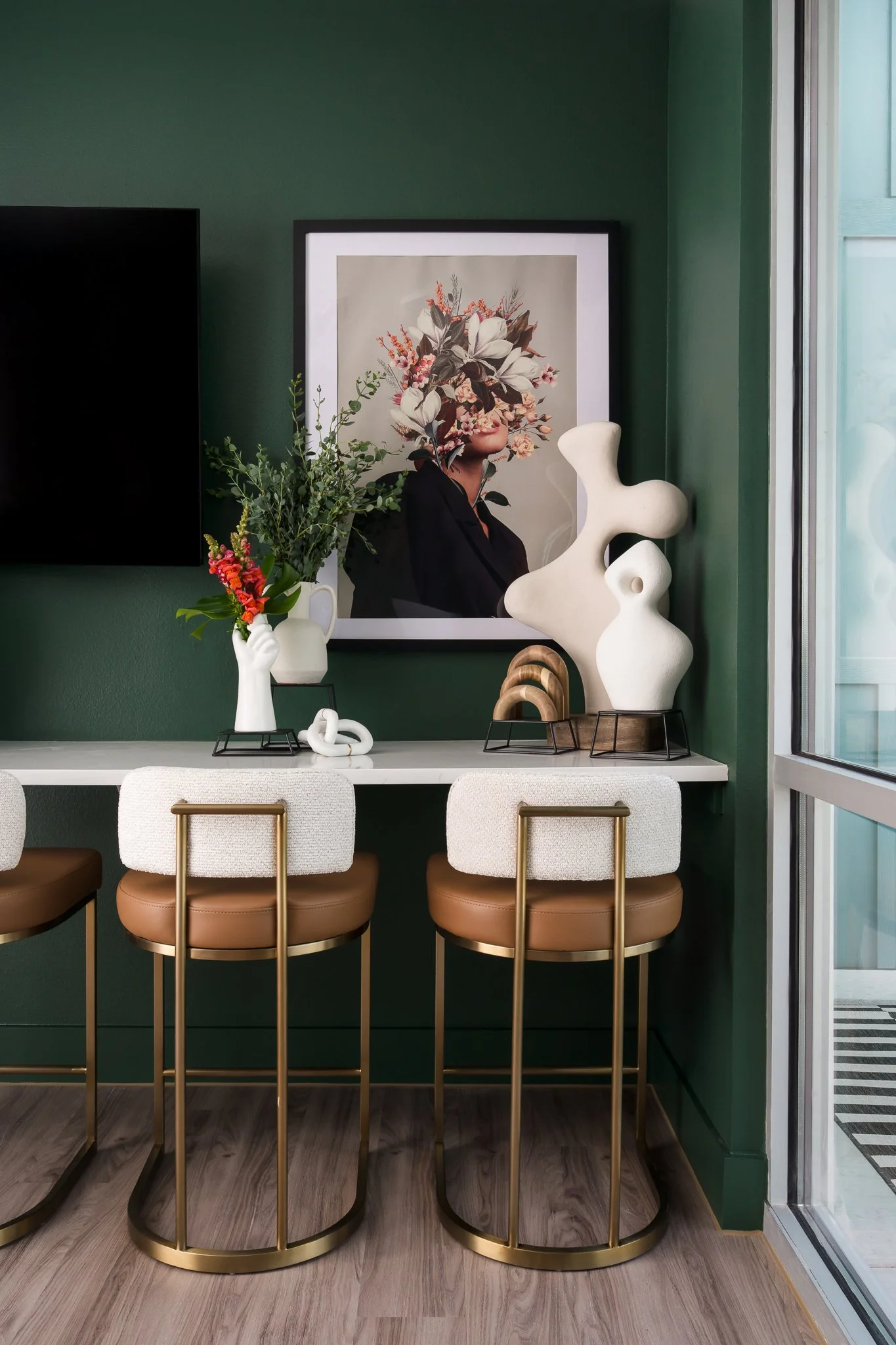 Interior of a modern room with a green wall, a white bar-height table, three stylish barstools with gold metallic frames and beige cushions, and decorative items including a vase with flowers, abstract sculptures, and a framed artwork of a person's f