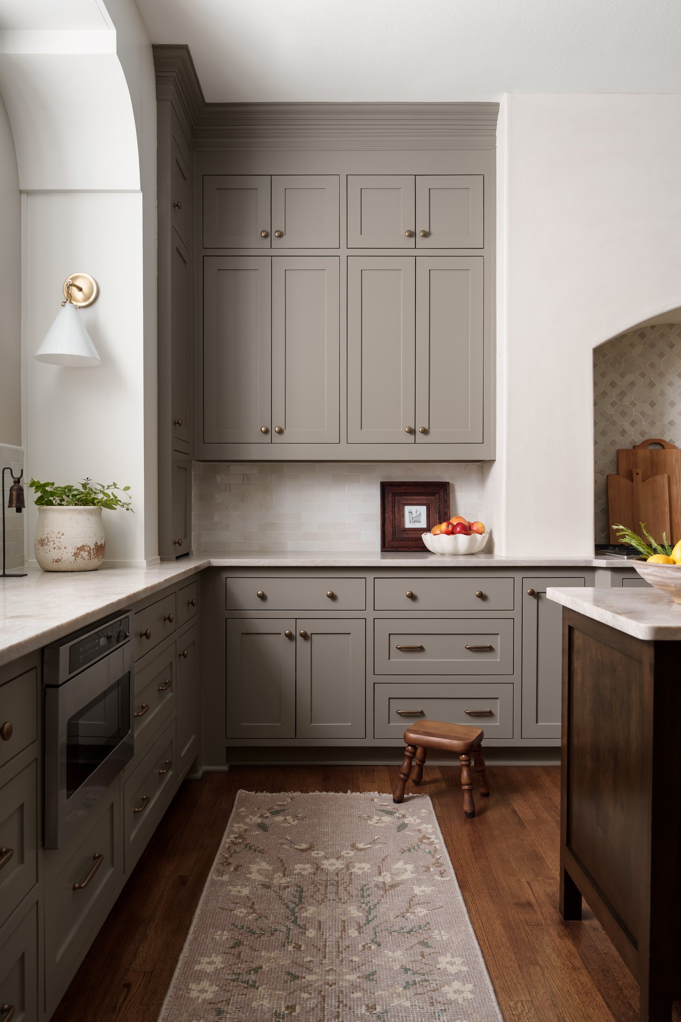 Kitchen with gray cabinets, white countertop, wooden stool, potted plant, bowl of apples, framed pictures, and a beige rug on wooden floor.