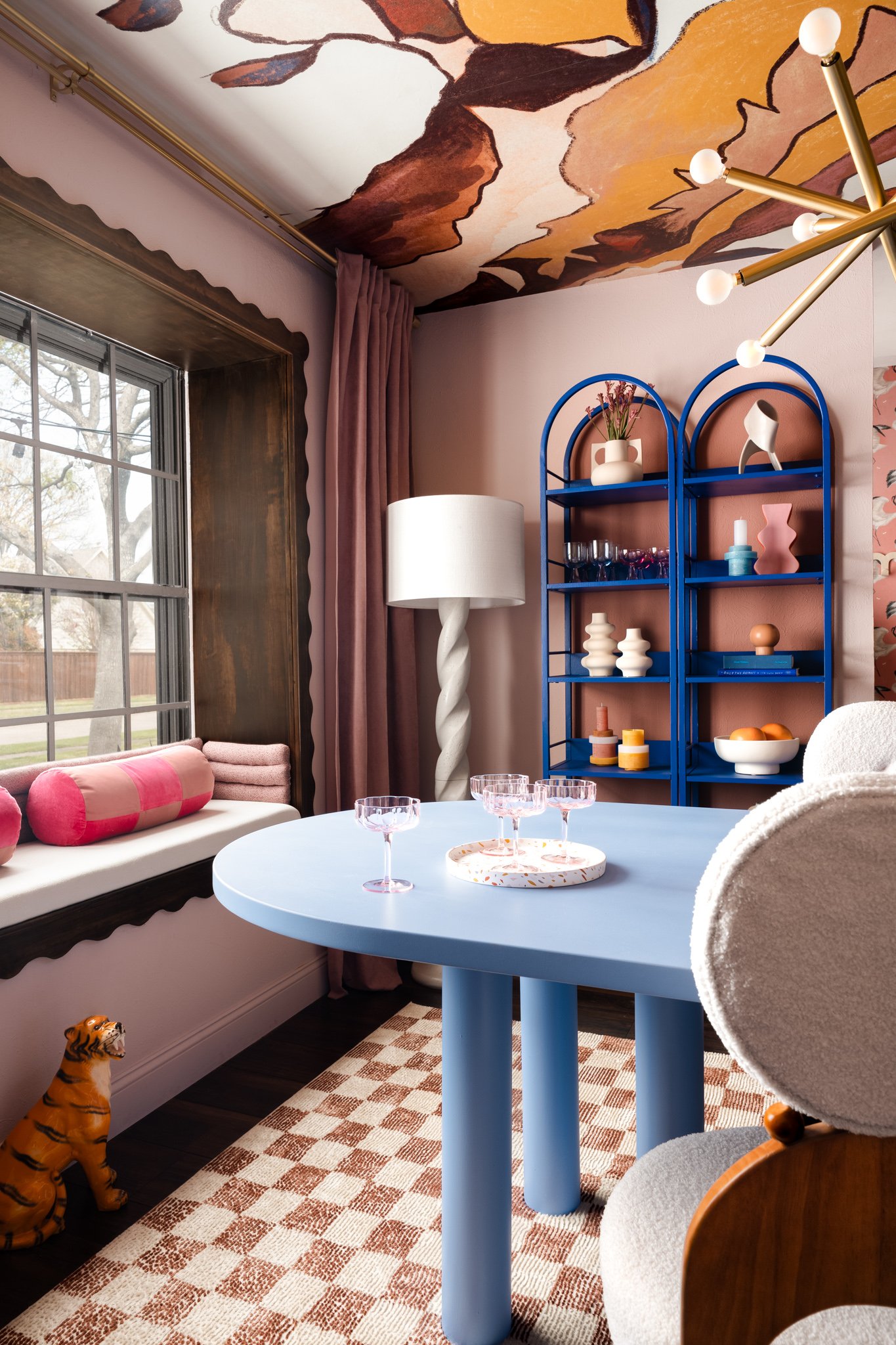 A cozy indoor scene with a pink and peach patterned wall, a window with wooden trim, a beige sofa with pink and peach pillows, a checkered rug, a round blue table, a white table lamp with a twisted base, a blue bookshelf with decorative vases and obj