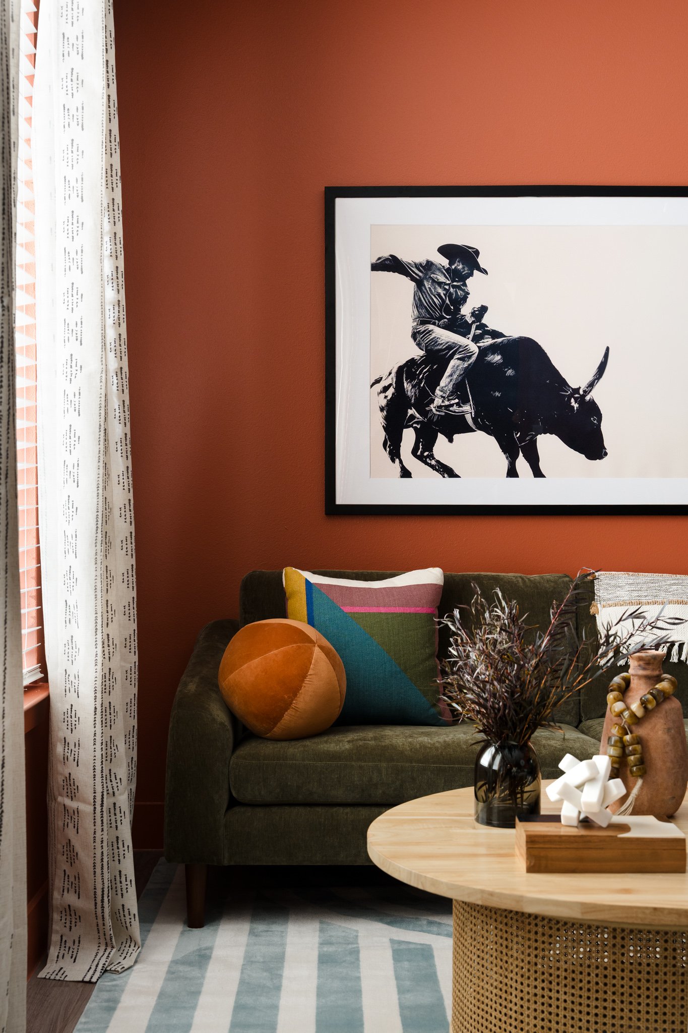 Living room with a green sofa, decorative pillows, a wooden coffee table with a black vase and dried flowers, a framed black-and-white image of a person riding a bull, and a patterned rug. Curtains hang by a window on the left, and various decorative