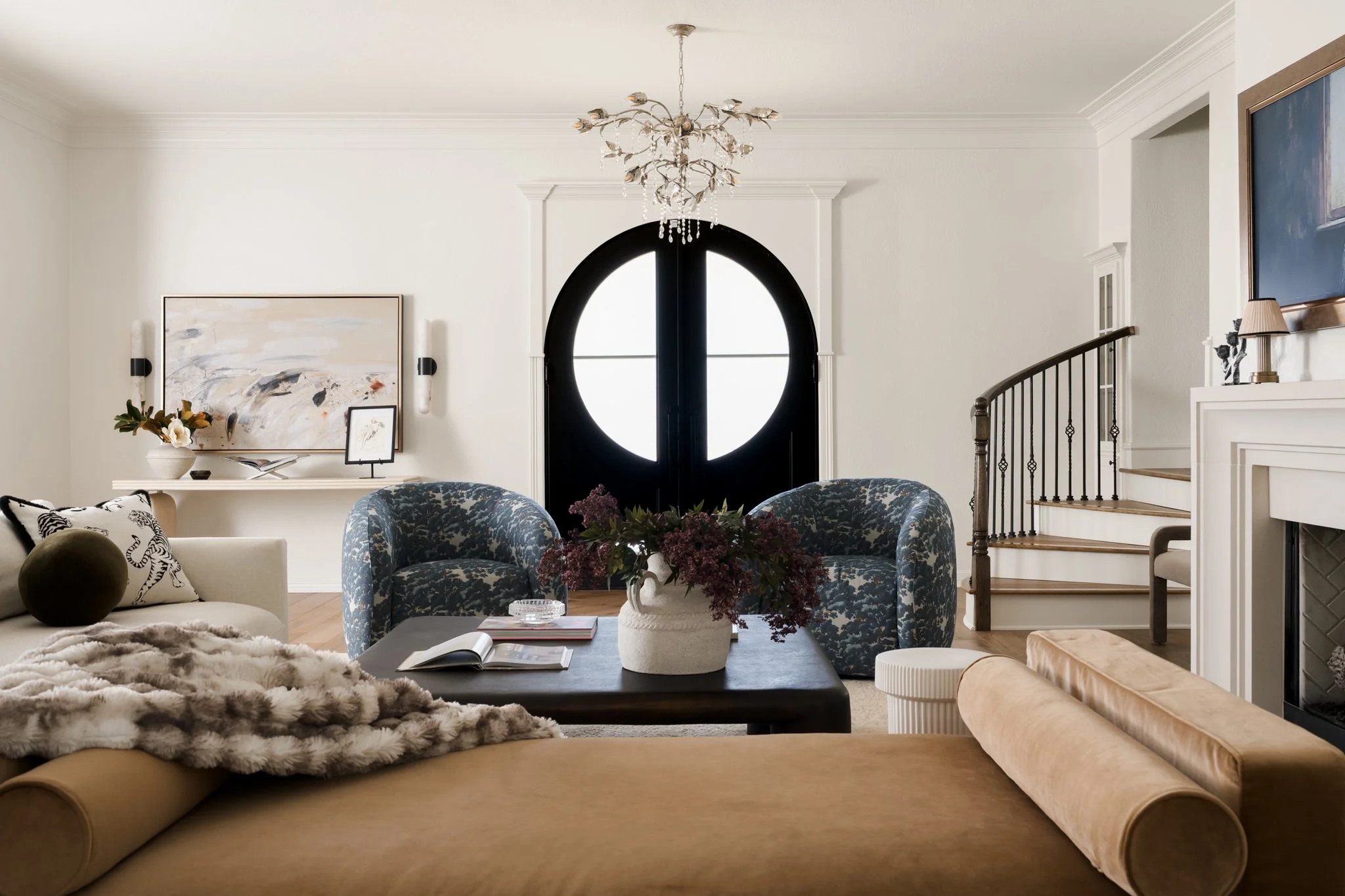 A cozy living room with a beige sofa topped with a fur throw, a black coffee table with an open book and a vase of flowers, two patterned armchairs, a round side table, a large abstract painting, a white console table with decorative items, a stairca