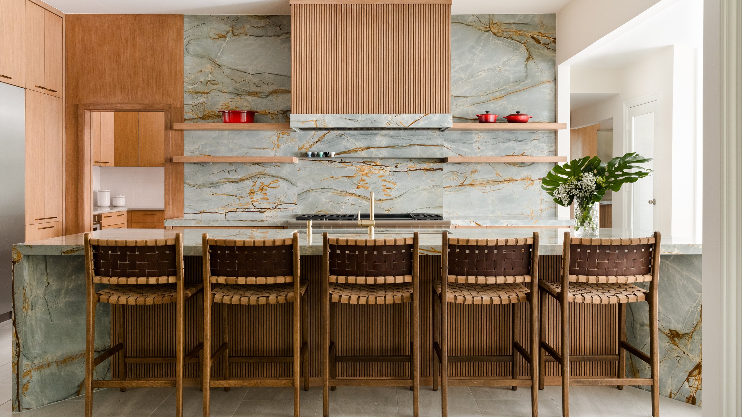 A modern kitchen with a marble backsplash, wooden cabinetry, a kitchen island with barstools, a stovetop, and a vase with large green leaves and white flowers.