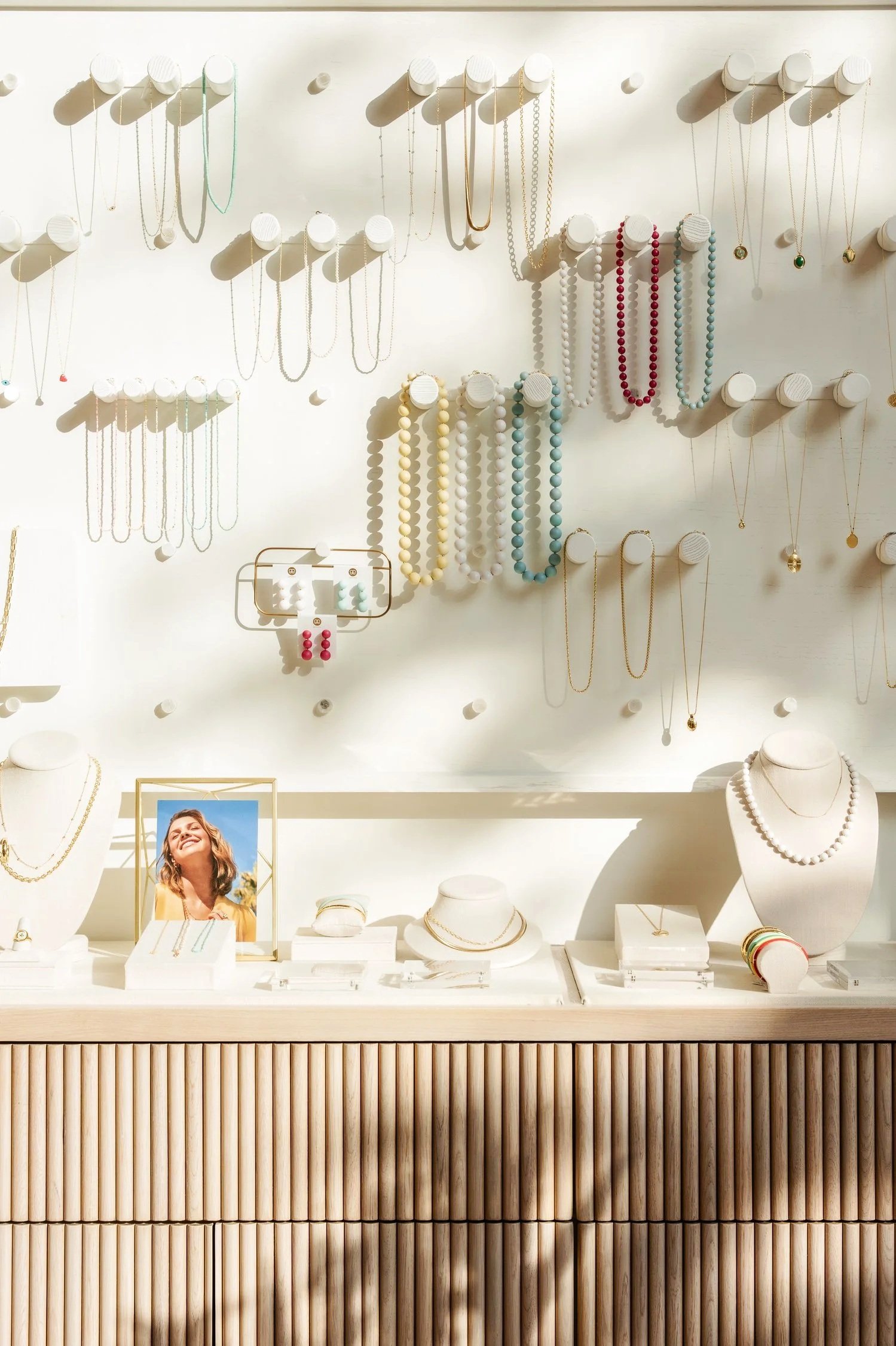 Jewelry display with necklaces, earrings, and bracelets on white stands and hooks, with a framed photo of a smiling woman on a white counter below.