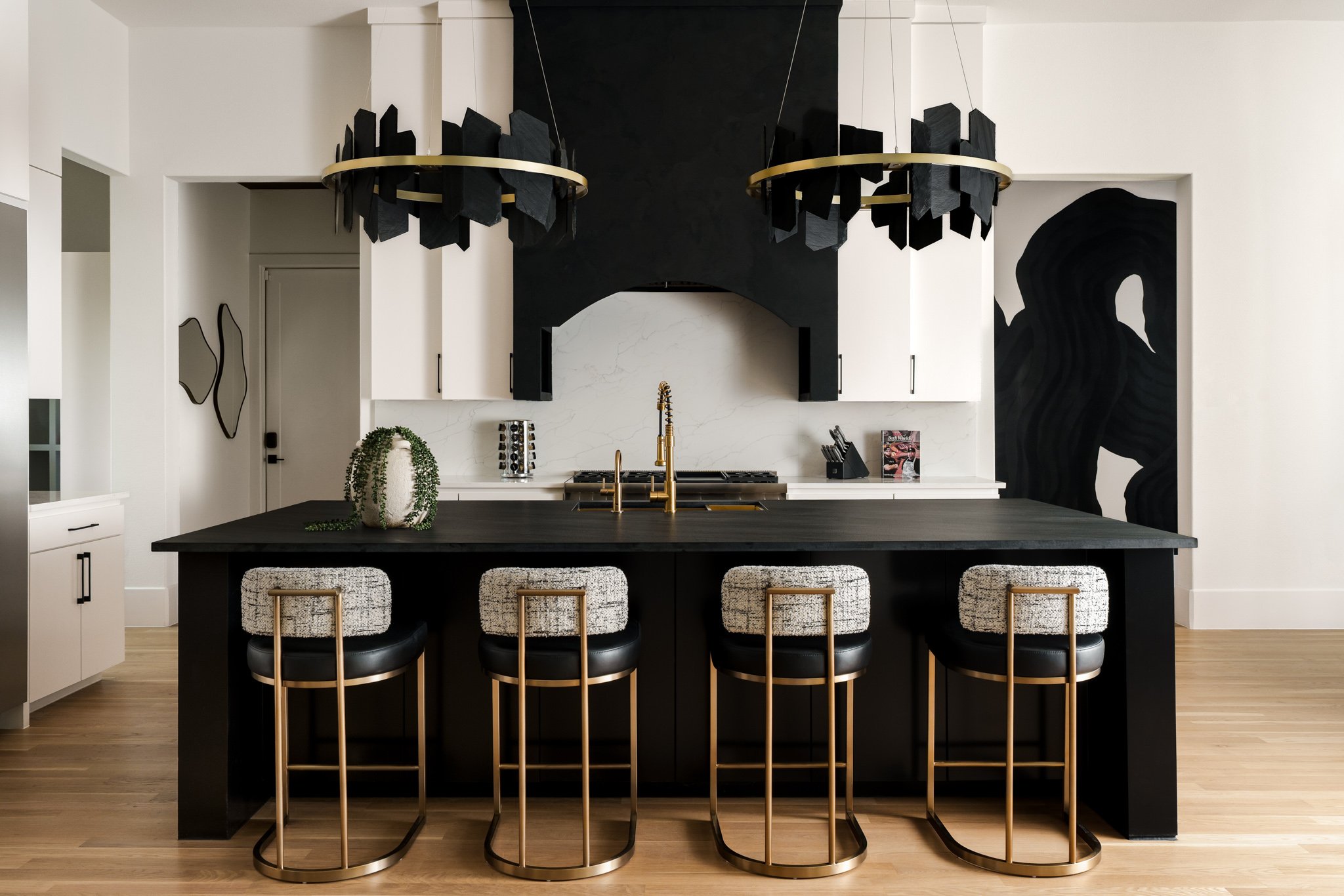 Modern kitchen with black island, white cabinets, black and white barstools with gold accents, black and gold chandeliers, and black wall art.