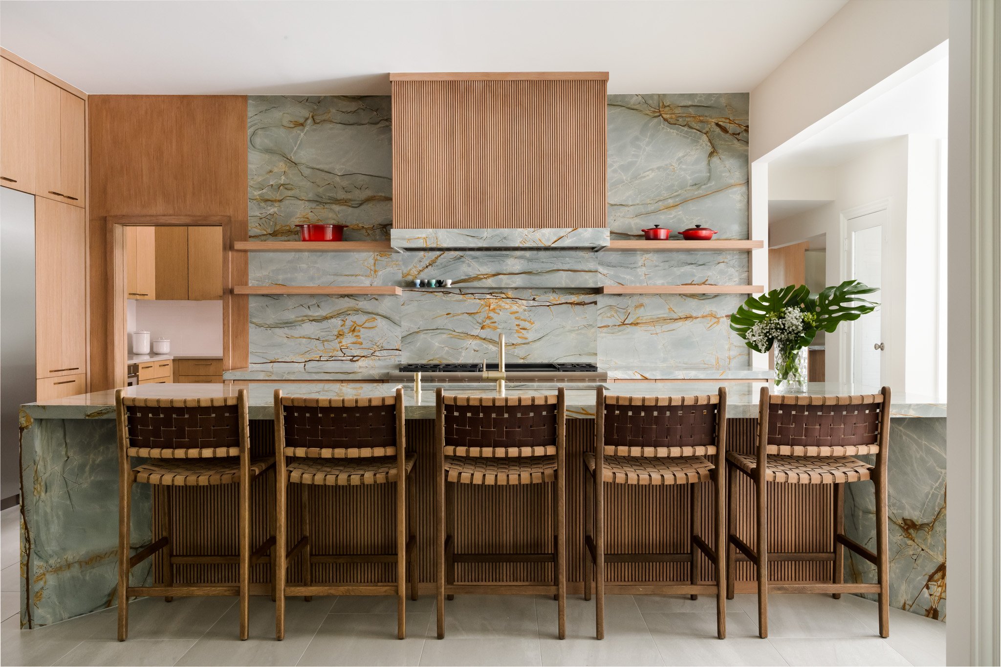 Modern kitchen with green marble backsplash and accents, a wooden panel over the range hood, a large island with seating, and a vase with white flowers and large green leaves.