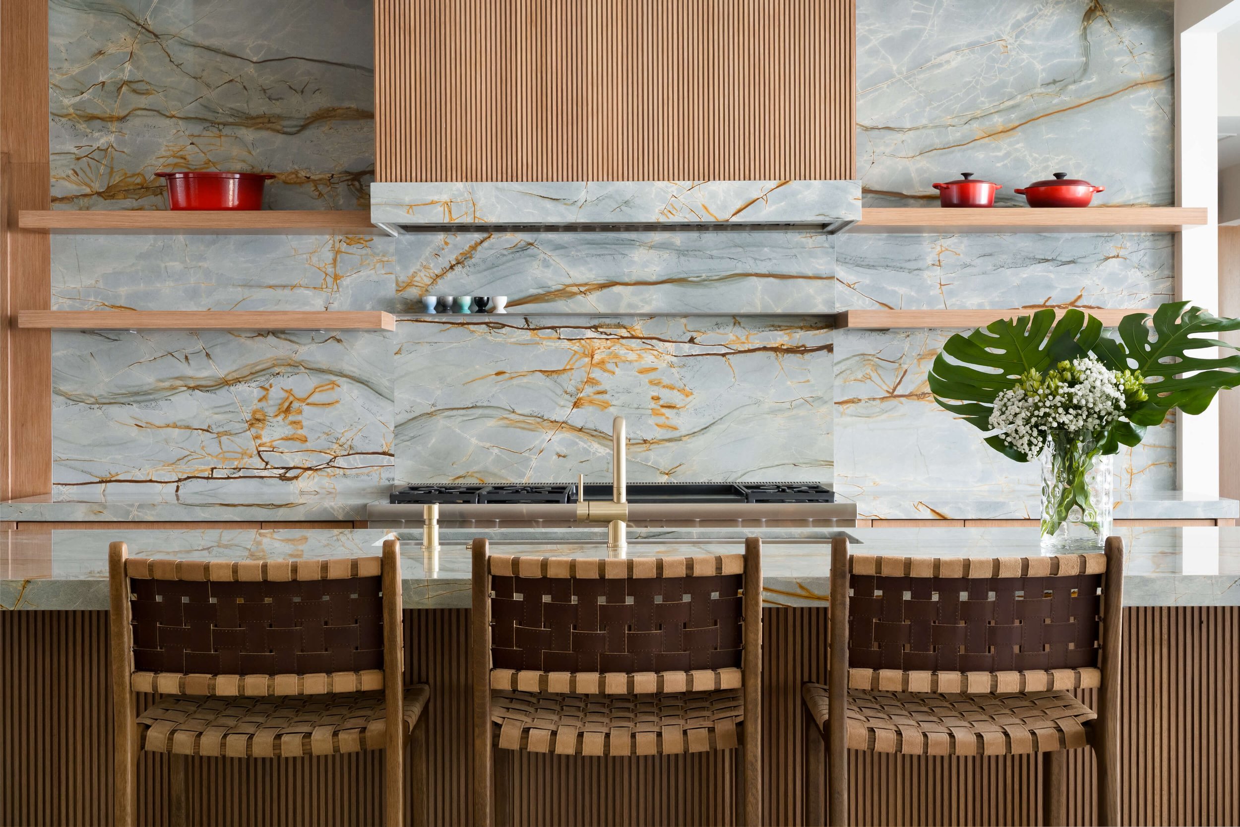 Modern kitchen with marble backsplash, wooden shelves, and black, white, and gray cups on the shelf. There is also a large green leafy plant with white flowers on the right side of the counter, and a wooden front row of chairs in the foreground.