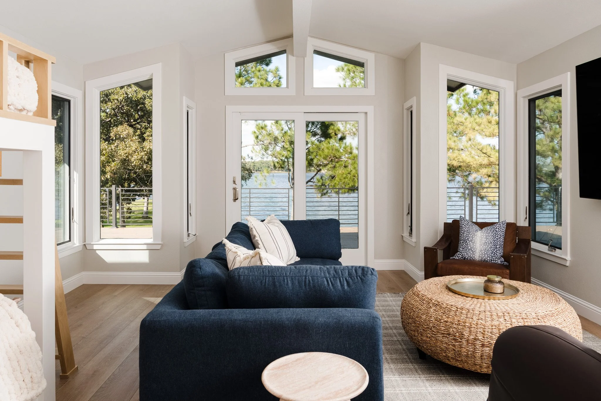 Living room with beige walls and multiple large windows overlooking greenery and water, with a navy blue sofa, a wicker ottoman, and a brown chair with a patterned pillow. Design by Lewis and Rose Interiors
