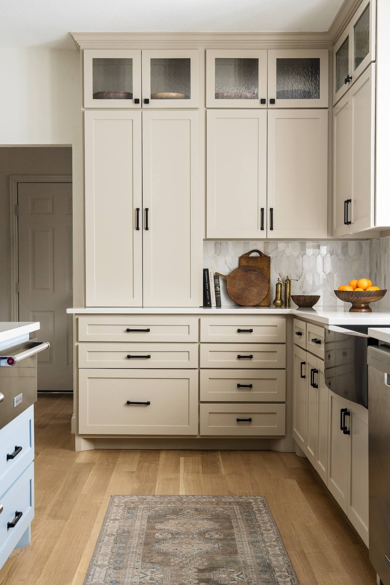 White kitchen cabinets with black handles in a modern kitchen. Countertop with decorative cutting boards, books, a small brass vase, a bowl, and a fruit bowl with oranges. Wooden floor and a small patterned rug.