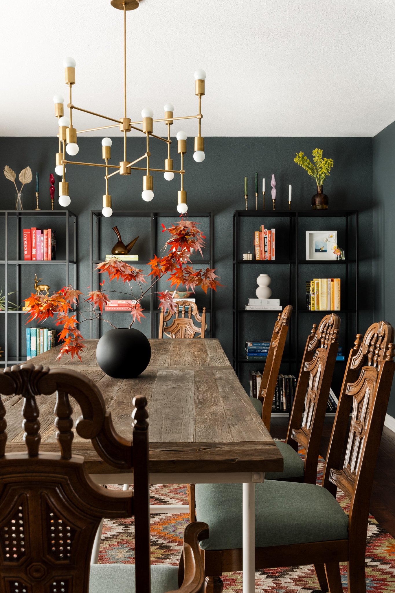 A dining room with a wooden table, chairs, black bookshelves, a modern gold chandelier, a vase with yellow flowers, and decorative items.