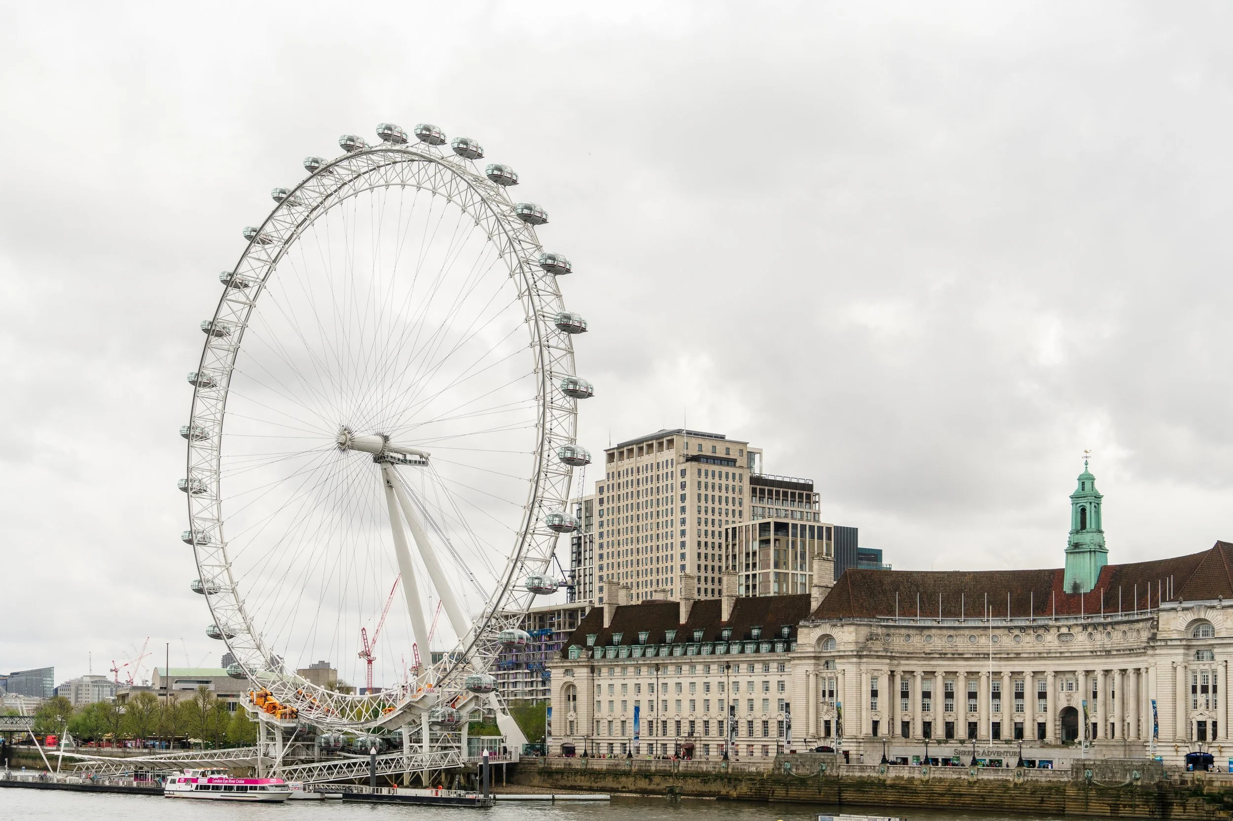 London Eye Ferris wheel on the River Thames with historic buildings in the background under cloudy sky.