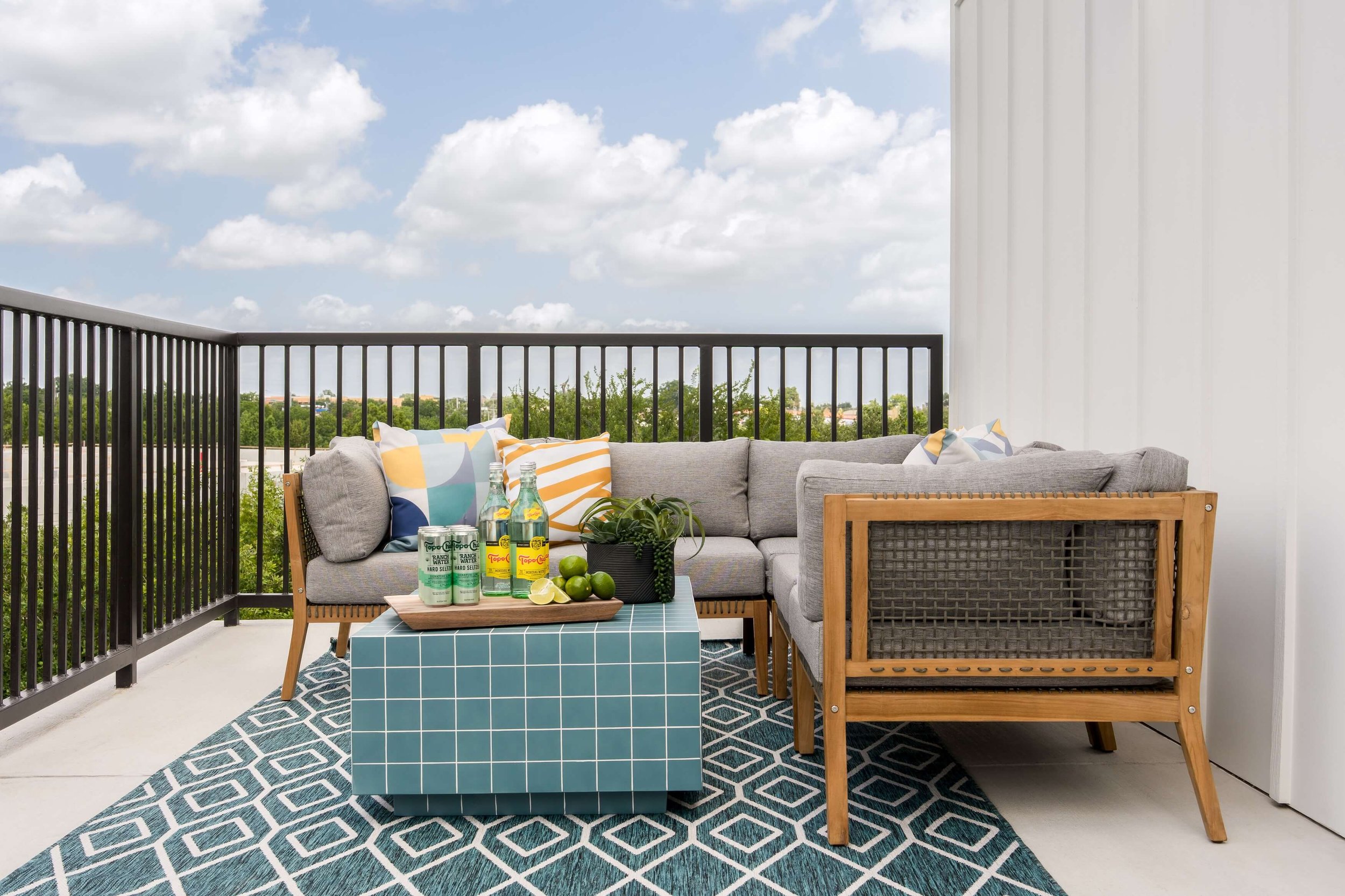 Outdoor balcony with a gray sectional sofa, colorful pillows, a blue tiled coffee table with drinks and limes, a potted plant, and a black railing, under a partly cloudy sky.