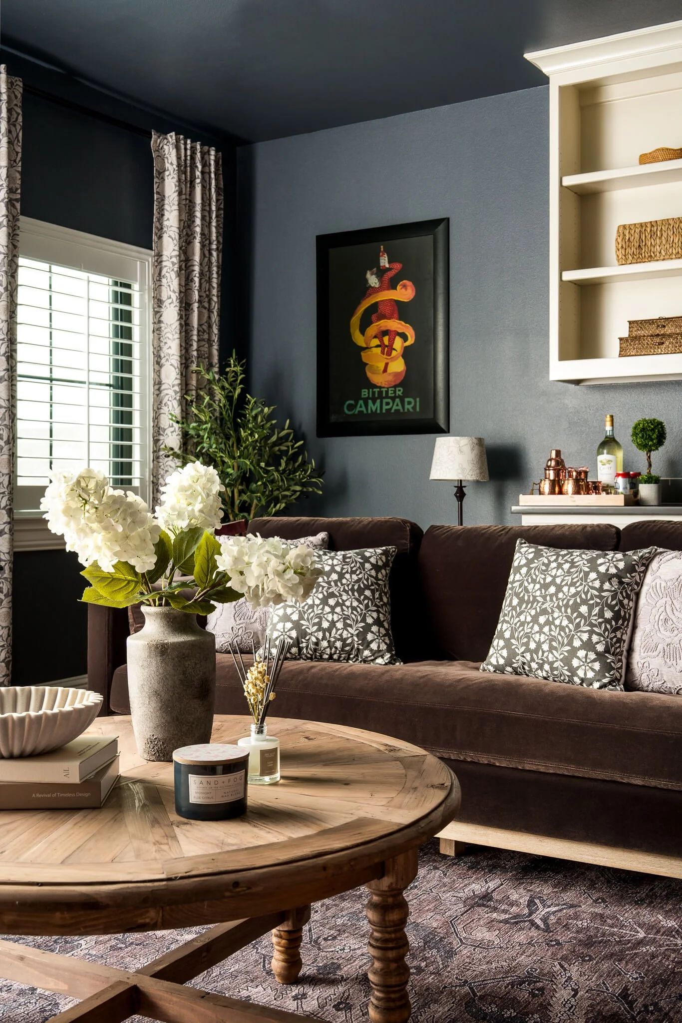 Living room with brown sofa, patterned pillows, wooden coffee table with candles, white flowers, and books, dark blue walls with a colorful vintage poster, window with white shutters and floral curtains, bookshelf with baskets, and a small lamp.
