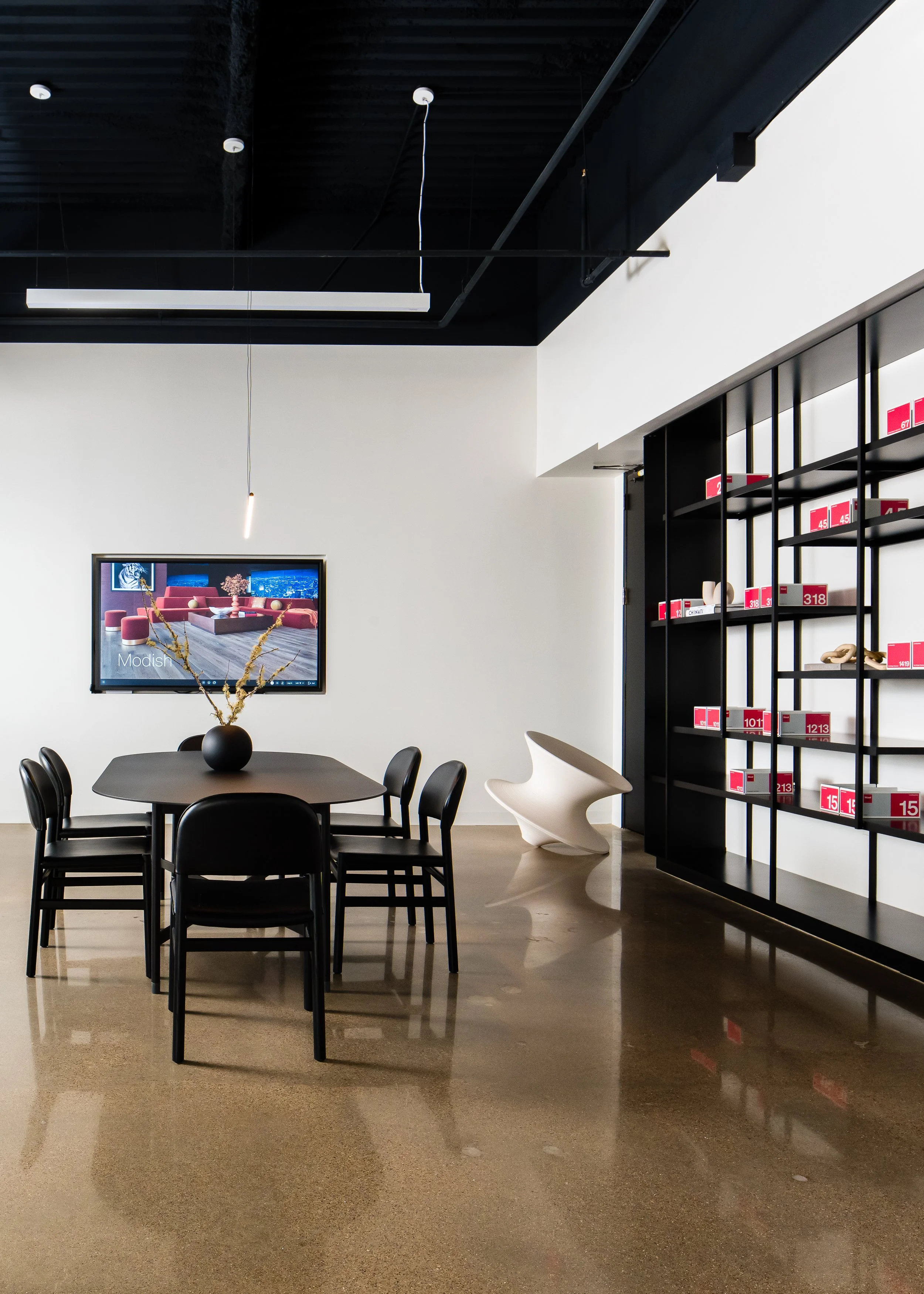 Modern interior space with a black table and chairs, a black bookshelf with red labels, a wall-mounted TV displaying furniture images, and a white lounge chair in the corner.