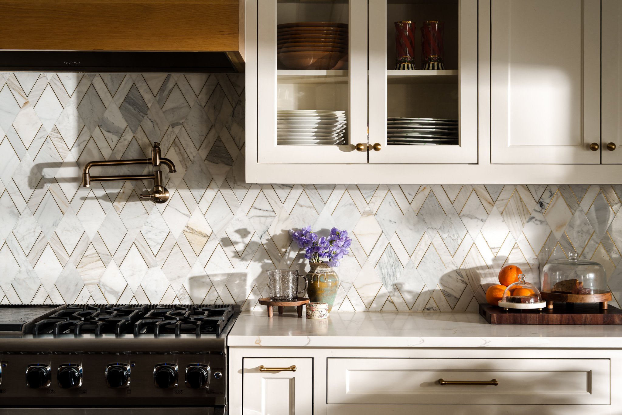 A modern kitchen with white cabinets, a marble backsplash, a gas stove, a gold faucet, and various decorative items including flowers, oranges, and a cake.