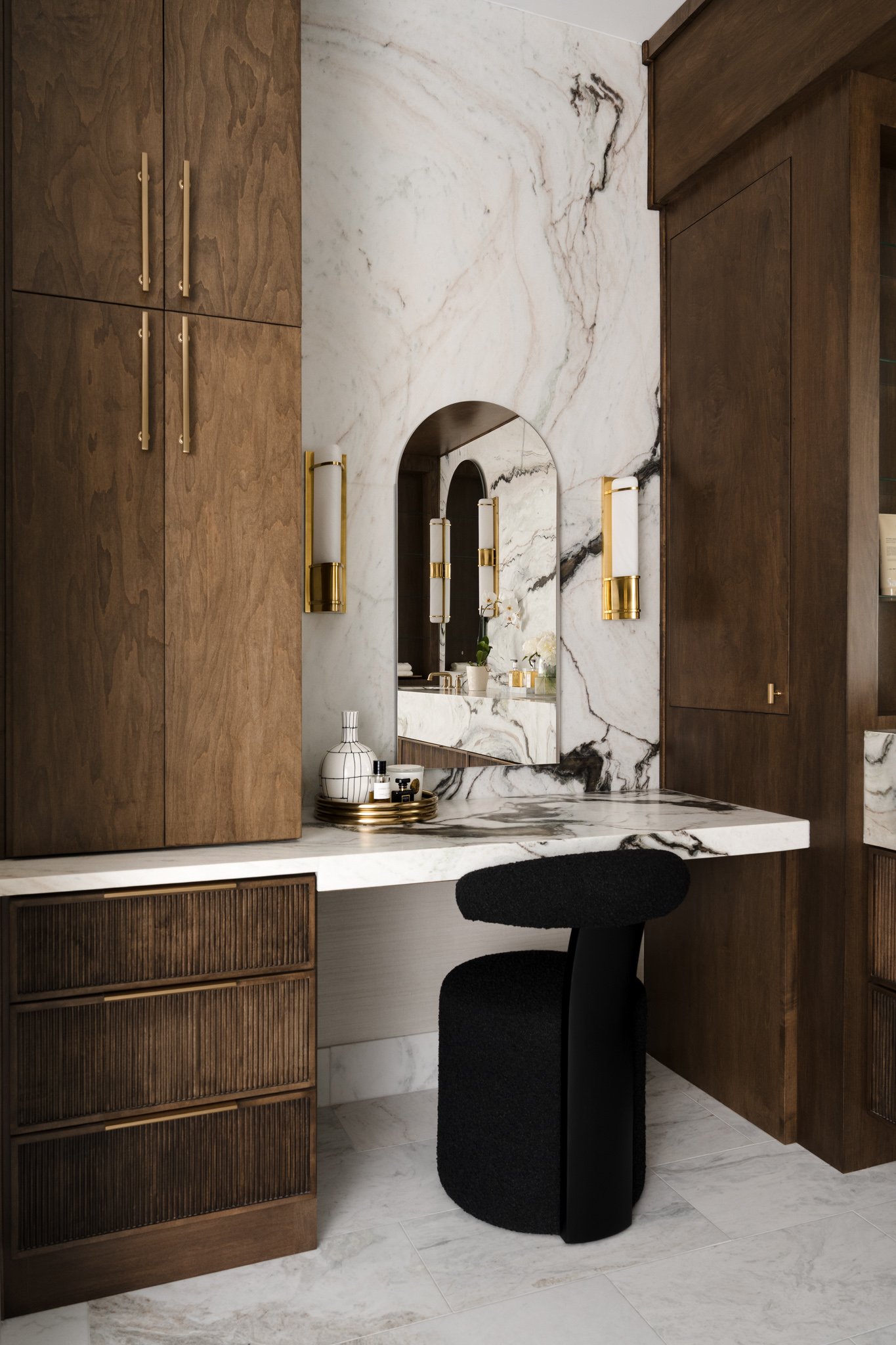 A modern vanity area with a marble countertop, dark wood cabinets, and a mirror. There are gold-accented wall sconces beside the mirror, small decorative items on the countertop, and a black stool at the vanity.