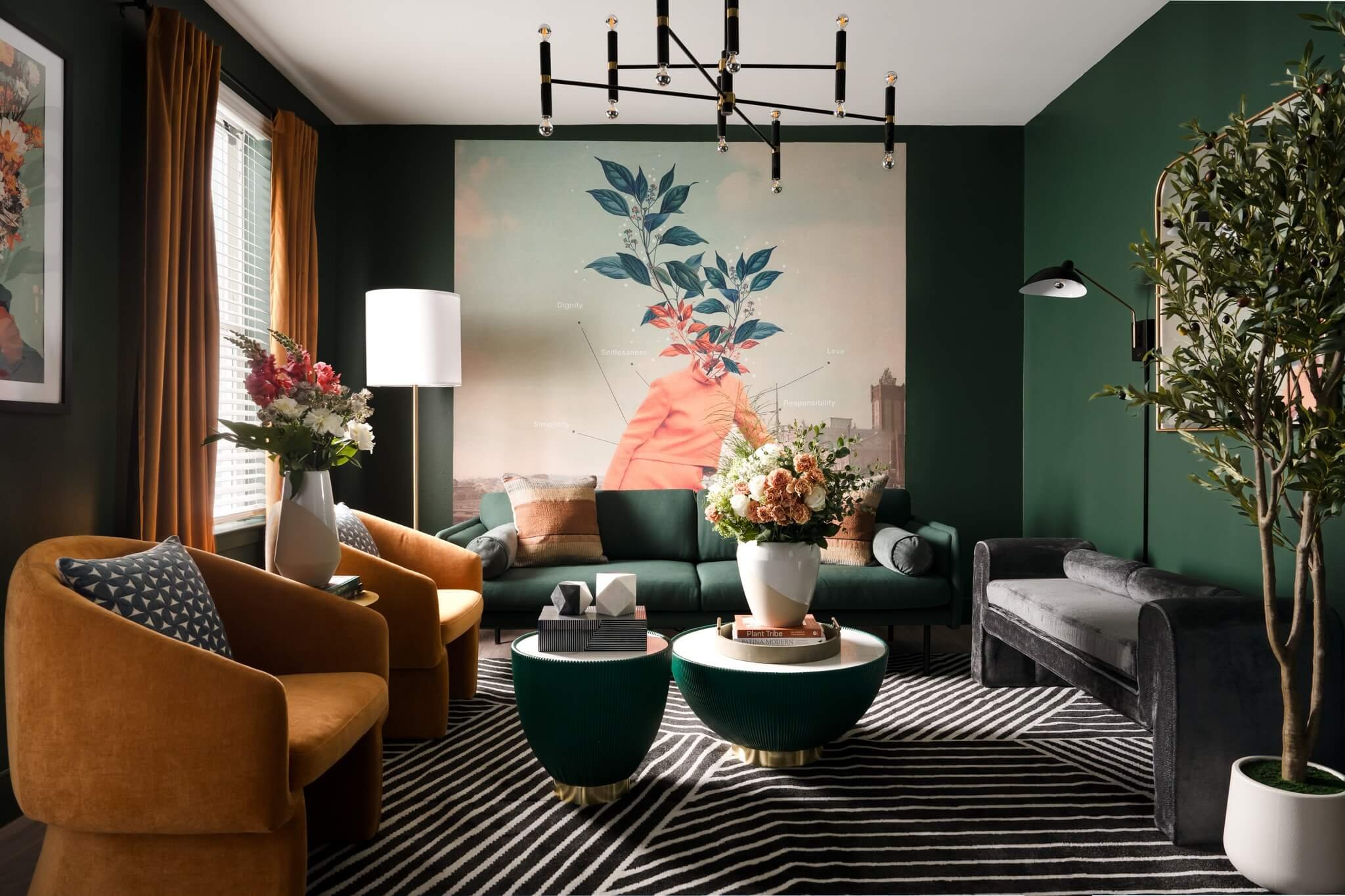 A modern living room with green walls featuring a large wall art of a person with a plant as their head. The room has a variety of colorful furniture including a teal sofa, orange armchair, gray velvet sofa, and a striped black and white rug. There a