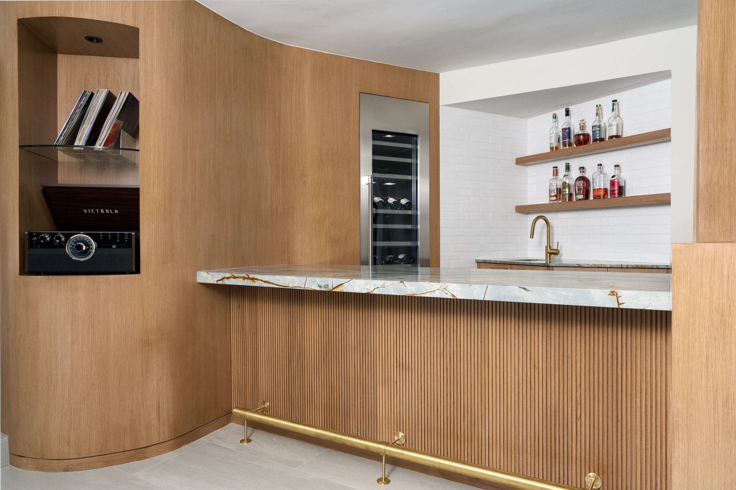 A modern kitchen with wood paneling, a marble countertop, a wine fridge, and shelves with liquor bottles.