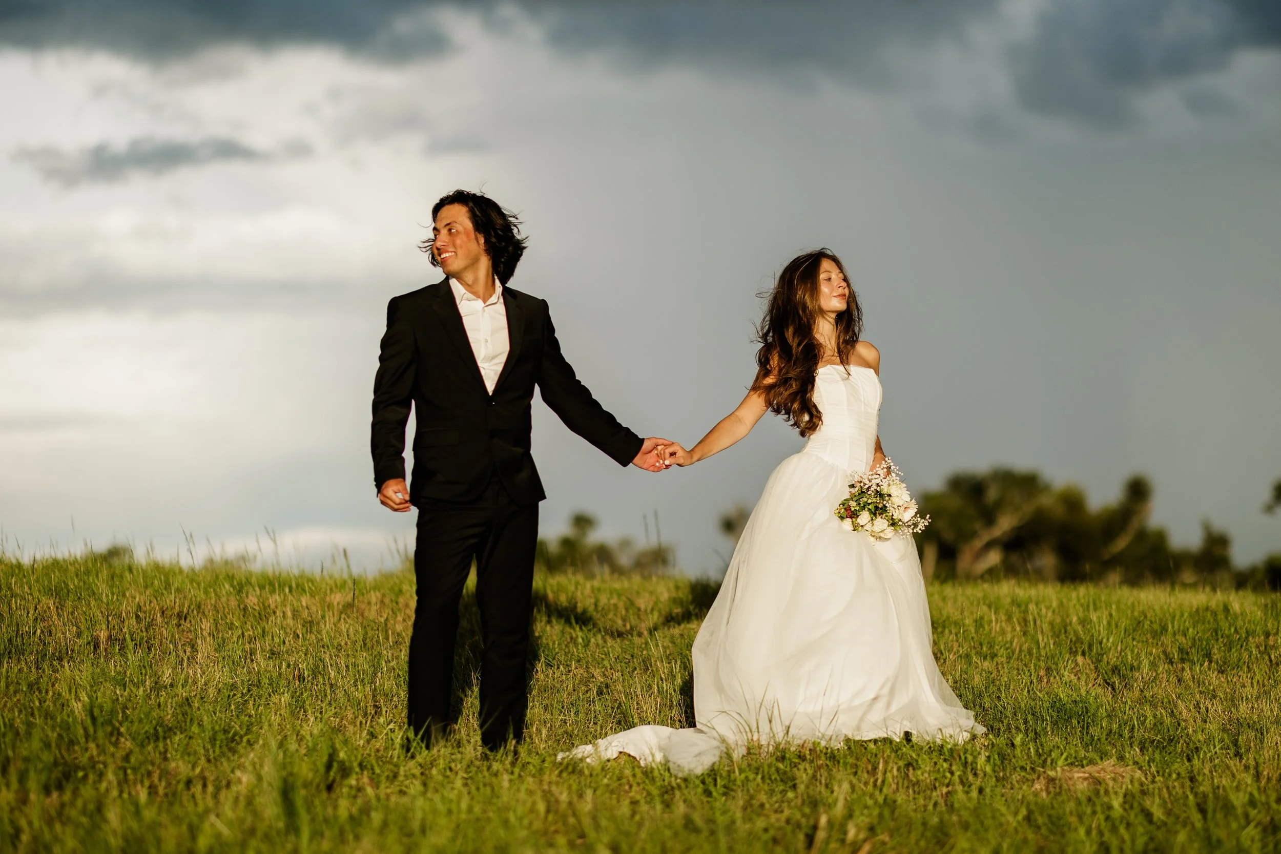 A couple dressed in wedding attire holding hands in an open field with a cloudy sky.