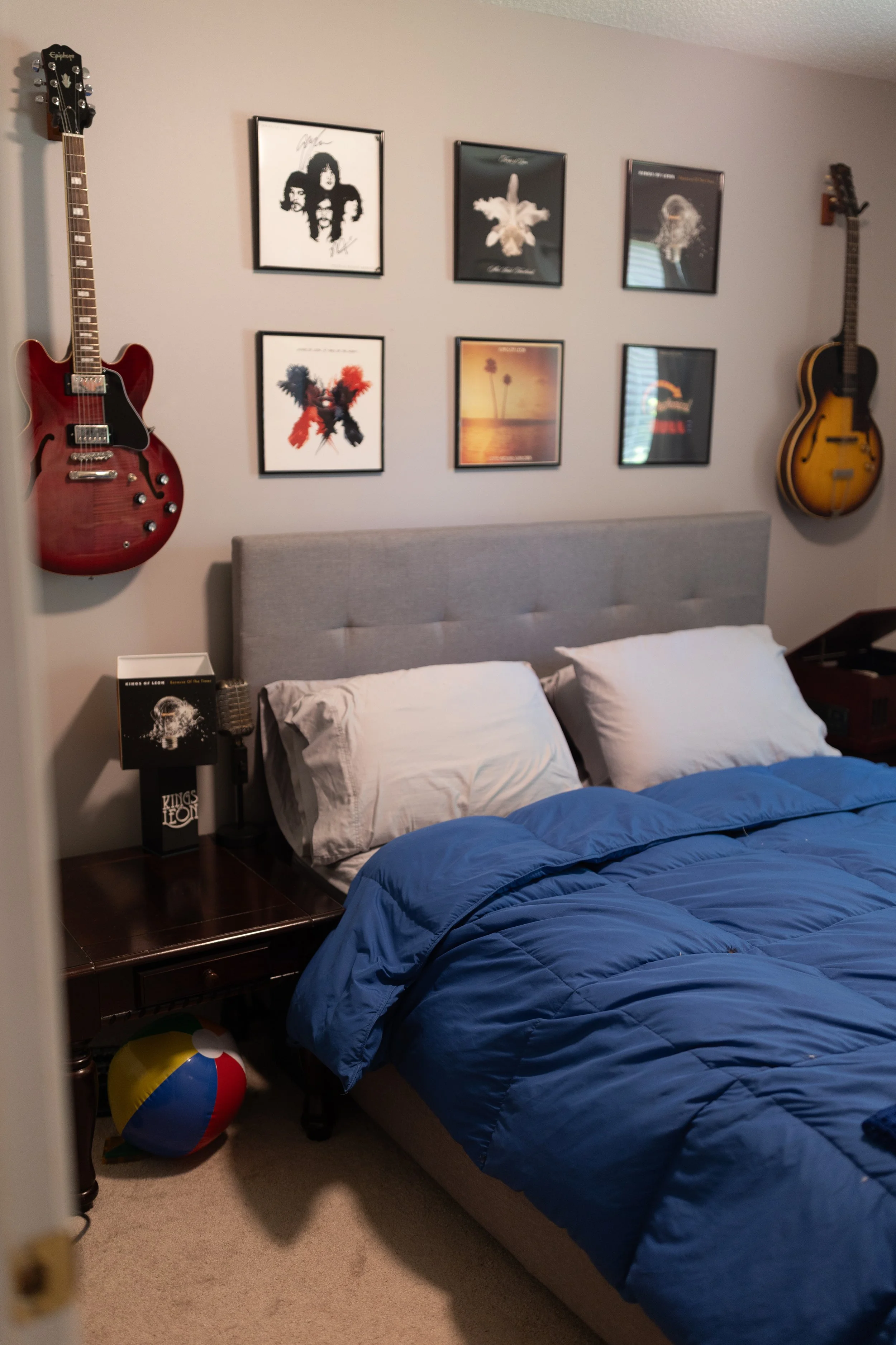 A bedroom with a bed covered in a blue comforter and white pillows. There are two guitars mounted on the wall, one on each side of the bed. Above the bed are six framed pictures, three on the top row and three on the bottom row, featuring artwork and