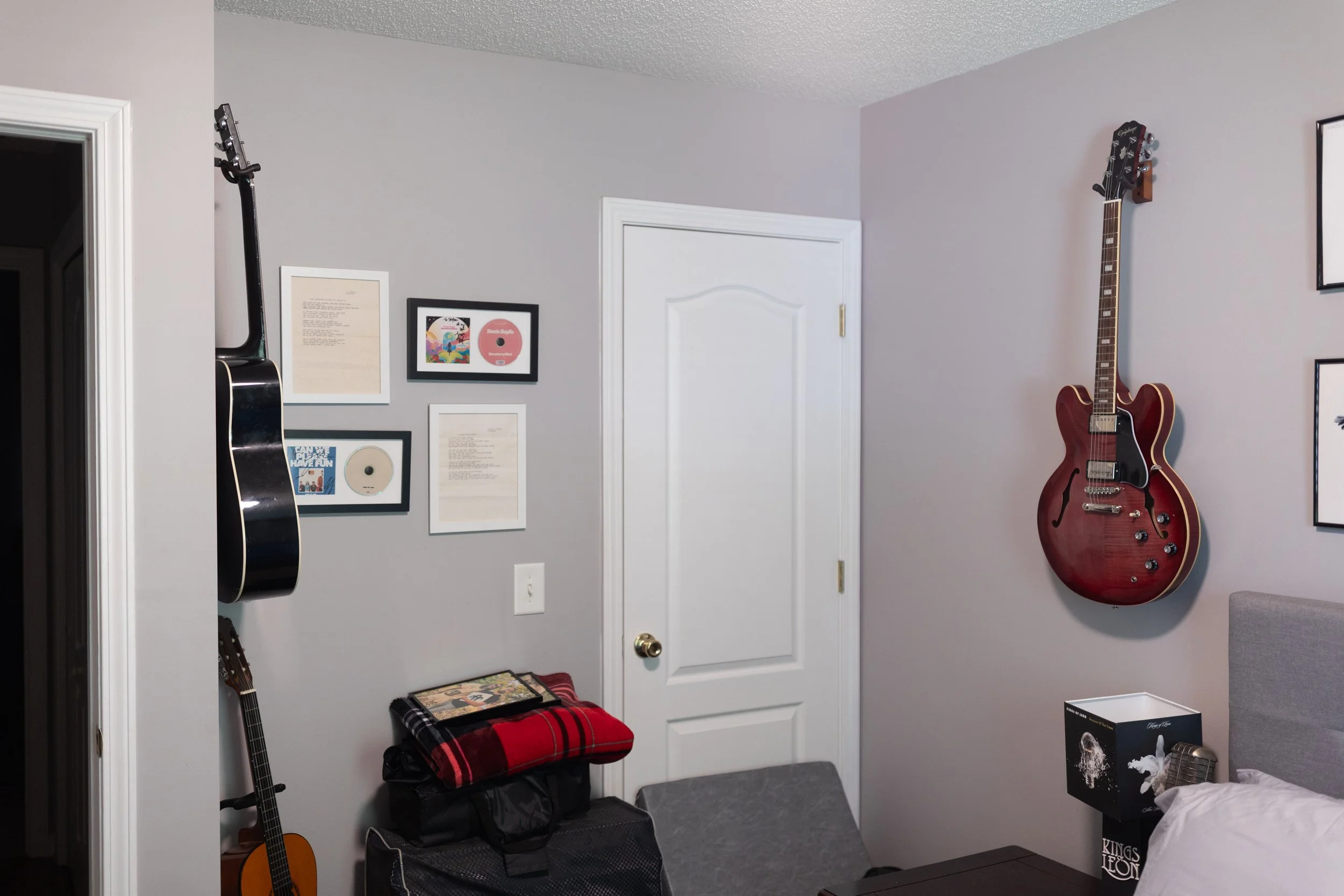 Room with guitars hanging on wall, framed pictures and albums, guitar on stand, plaid blanket on small chair, books and boxes on floor.