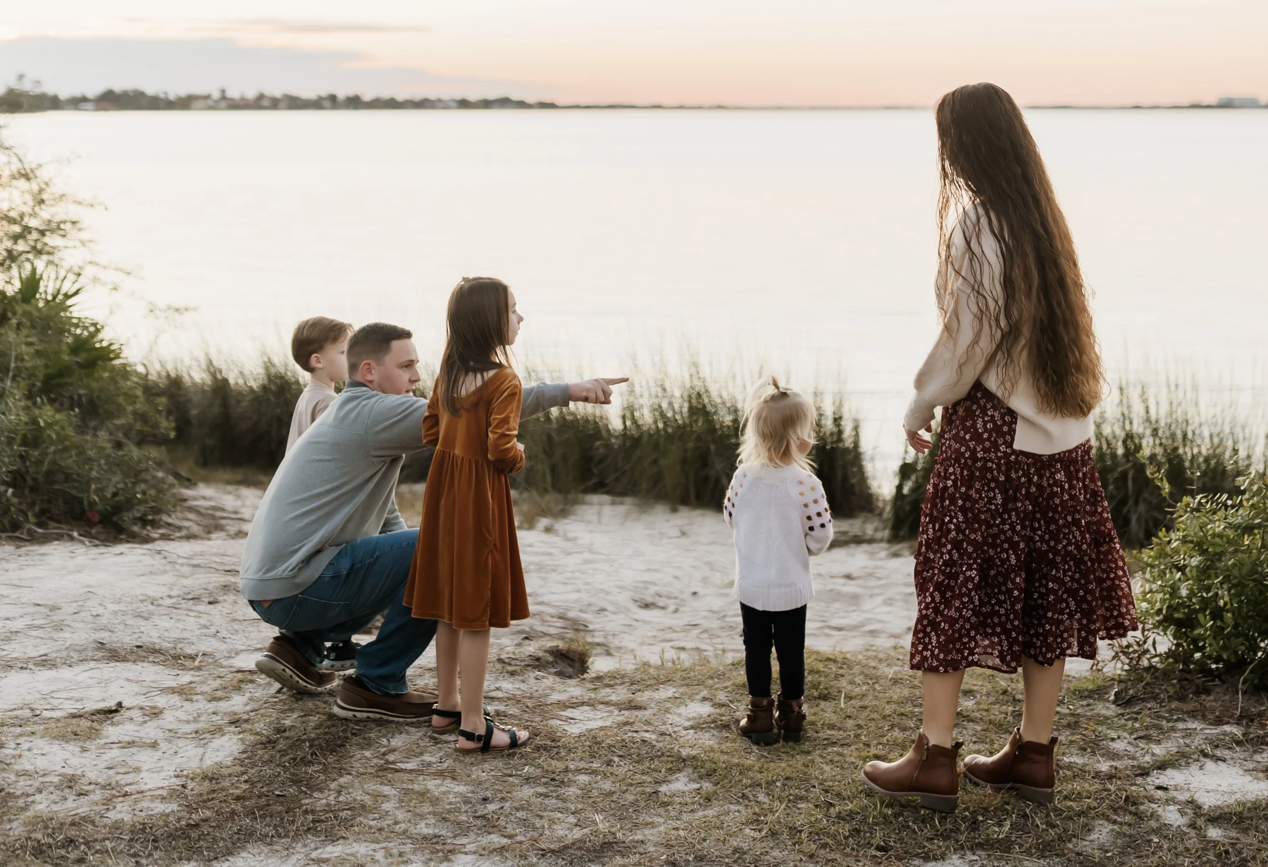 A woman with long, wavy hair standing on a sandy shore facing a group of four children, in front of a body of water at sunset.